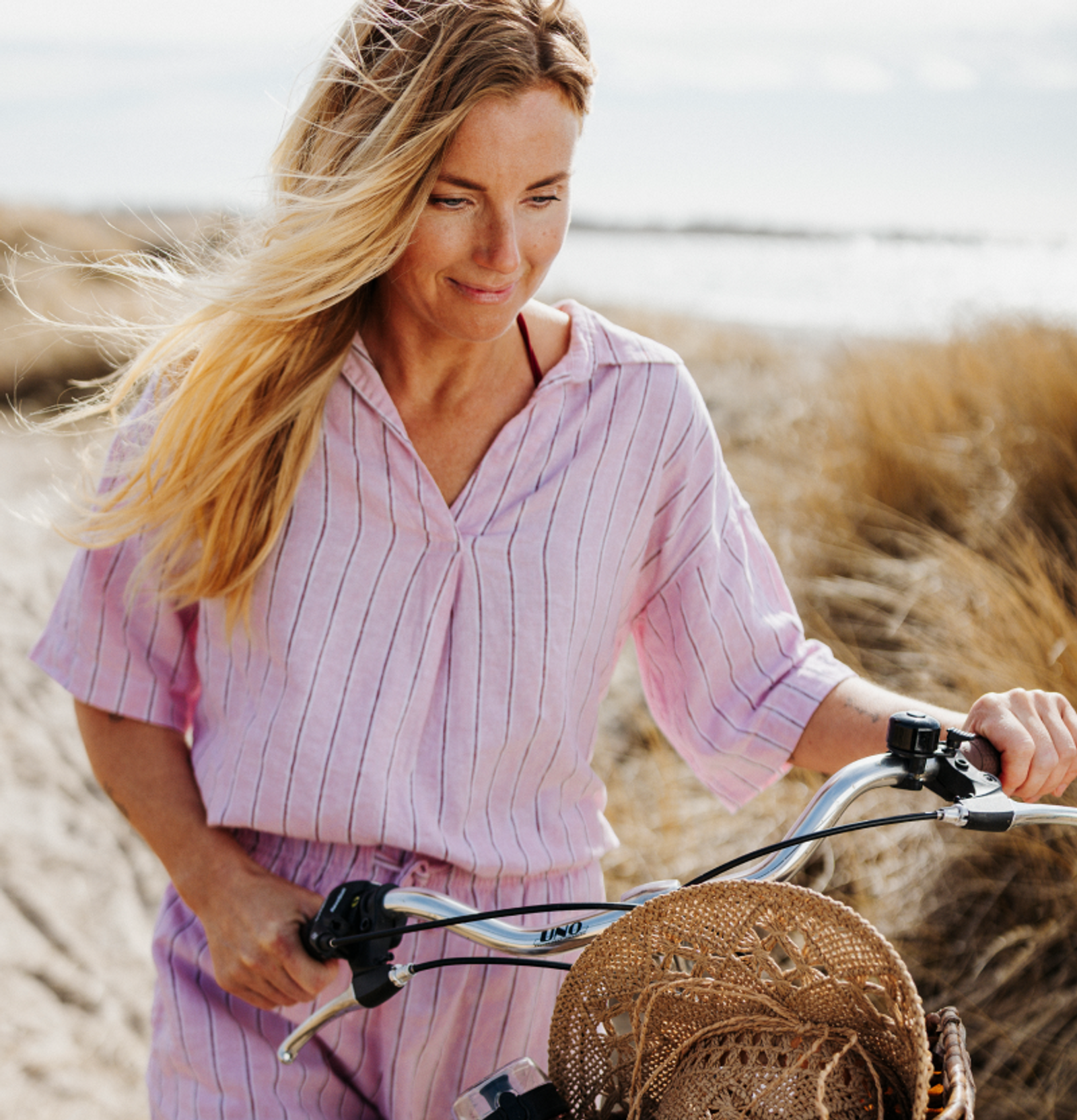 A blonde woman in a pink striped outfit walks a bicycle on a sandy path, a straw hat in the bike's basket.