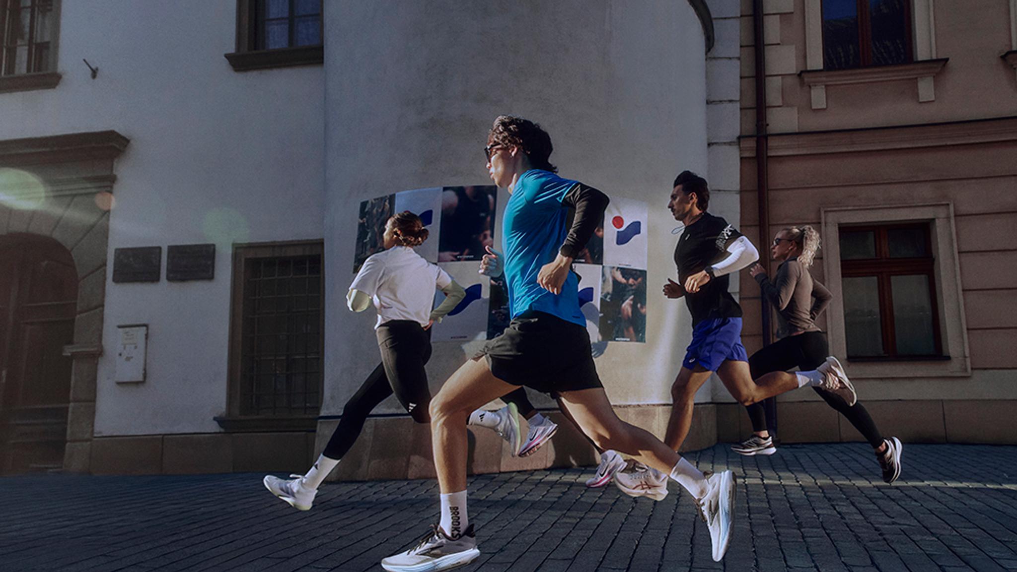 Four people in athletic wear run on a cobblestone street past old buildings on a sunny day.