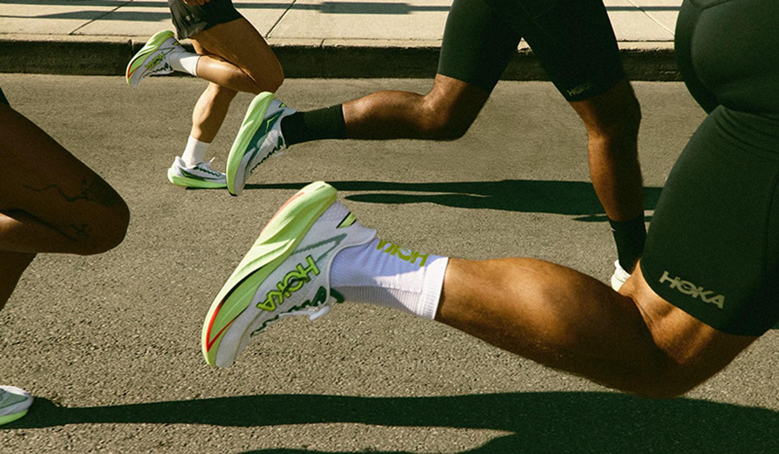 Close-up of multiple runners' legs and Hoka shoes in white, green, and neon yellow as they run on pavement.