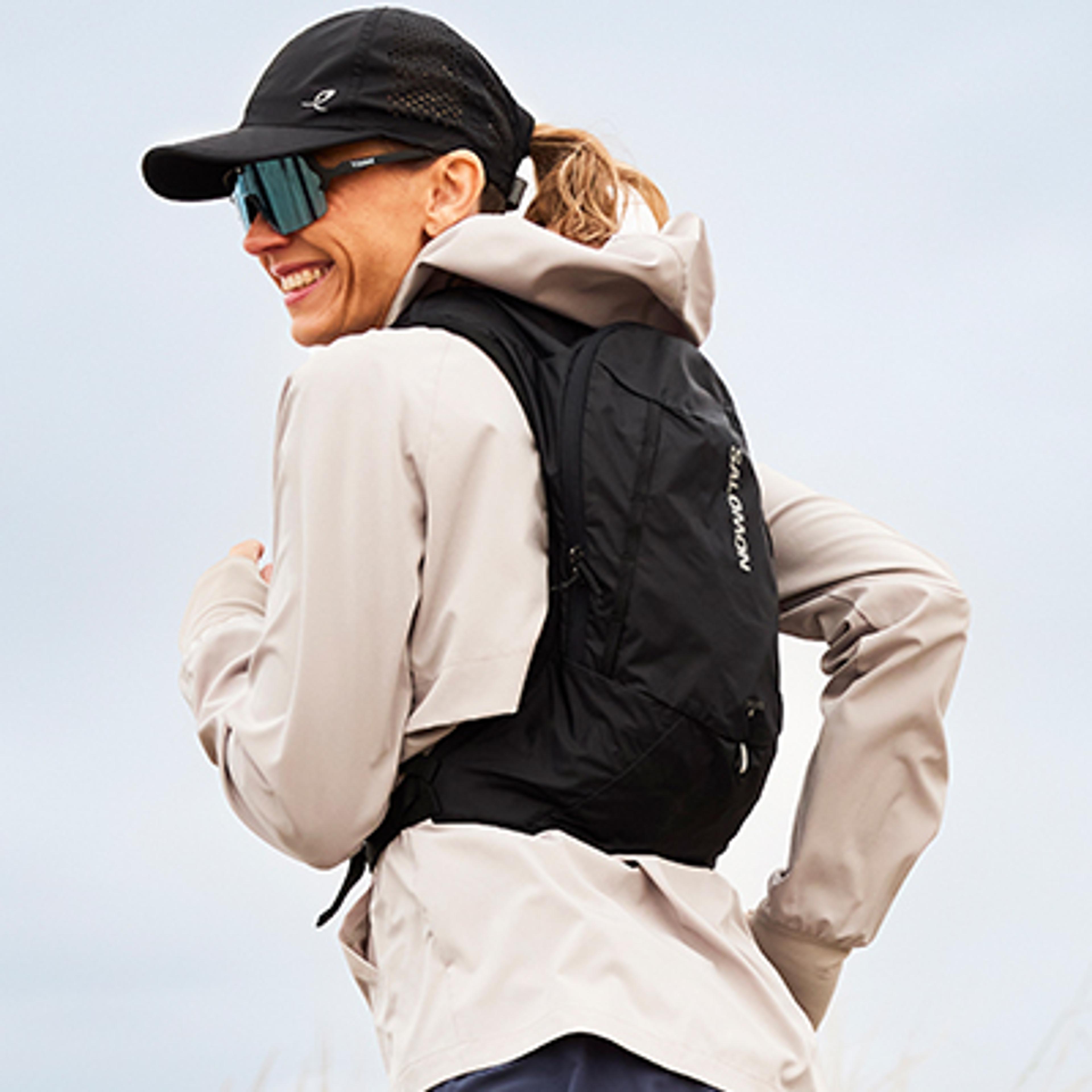Smiling woman in a black cap, turquoise sunglasses, beige jacket, and black Salomon running pack.