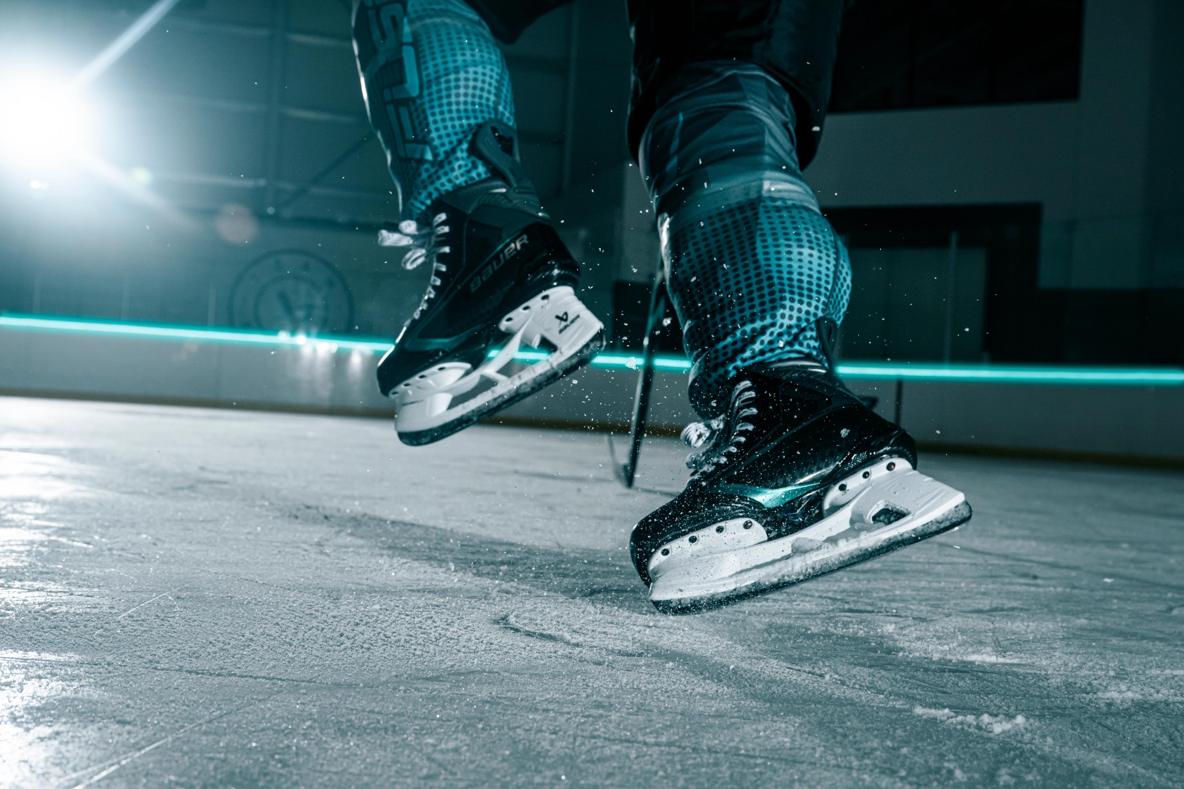 A person wearing black ice skates on an ice rink, with one skate lifted slightly off the ice.