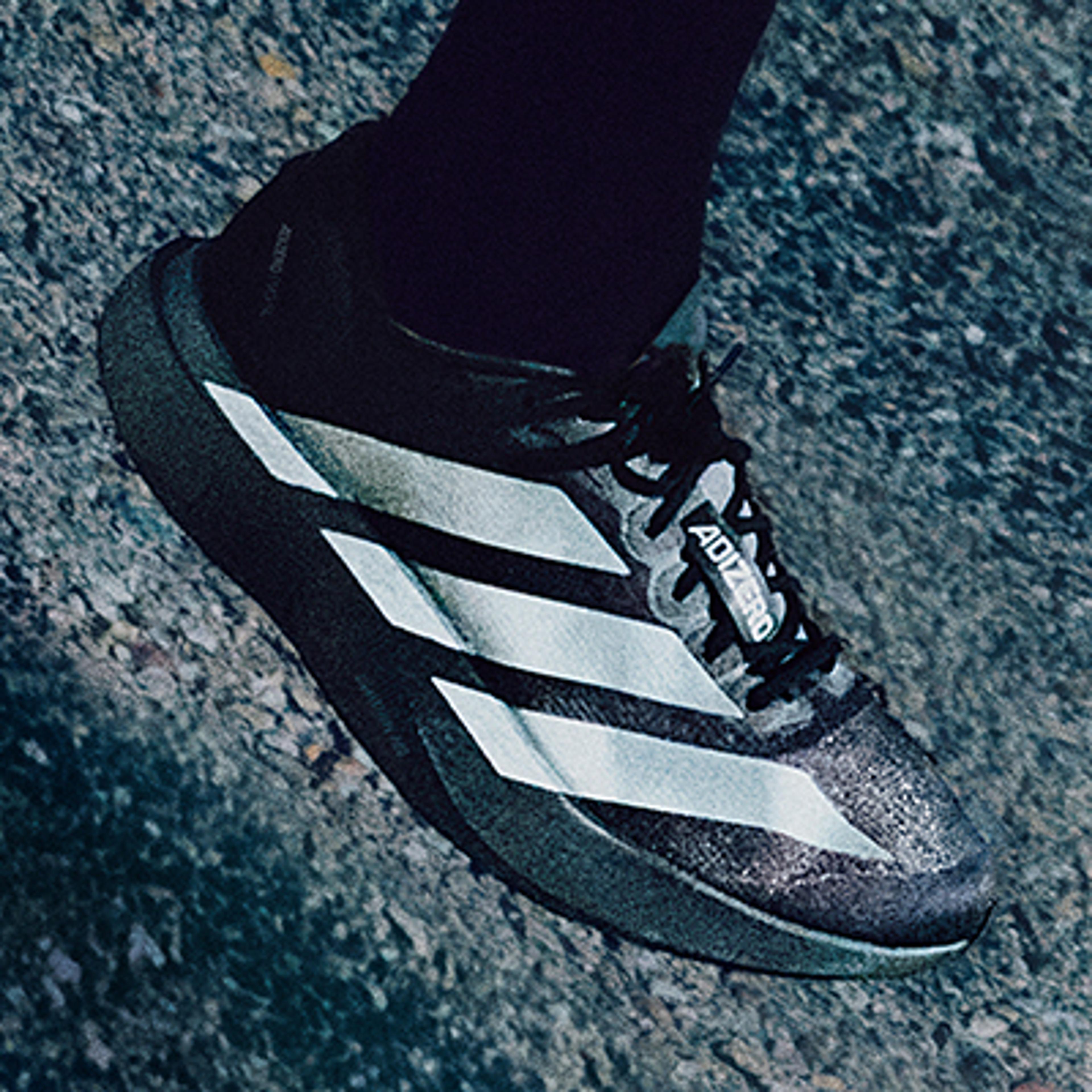 A white Adizero running shoe with blue and green stripes on a foot wearing a black sock.