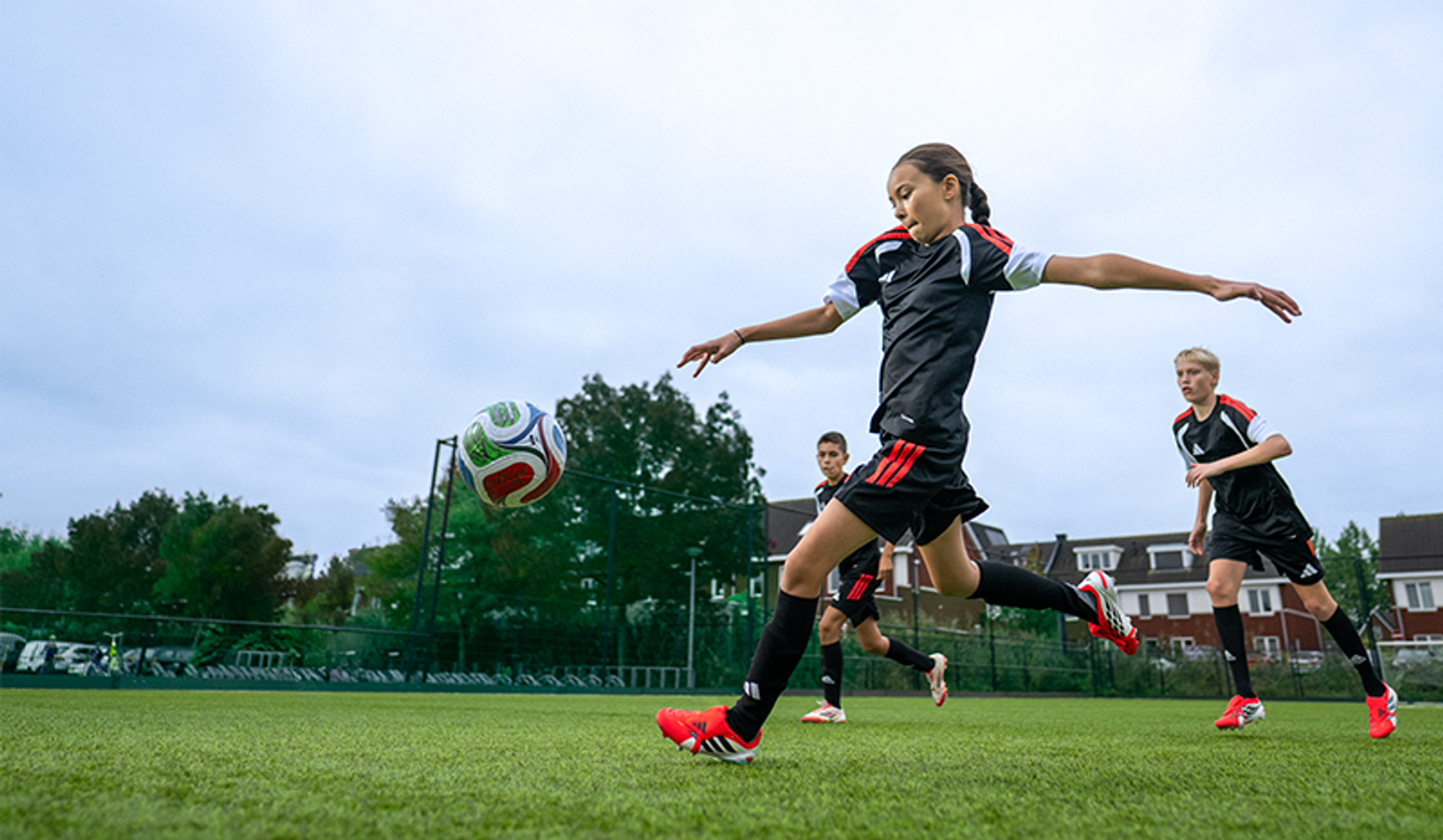 A girl in a black and red soccer uniform kicks a ball mid-air on a green field, with teammates nearby.