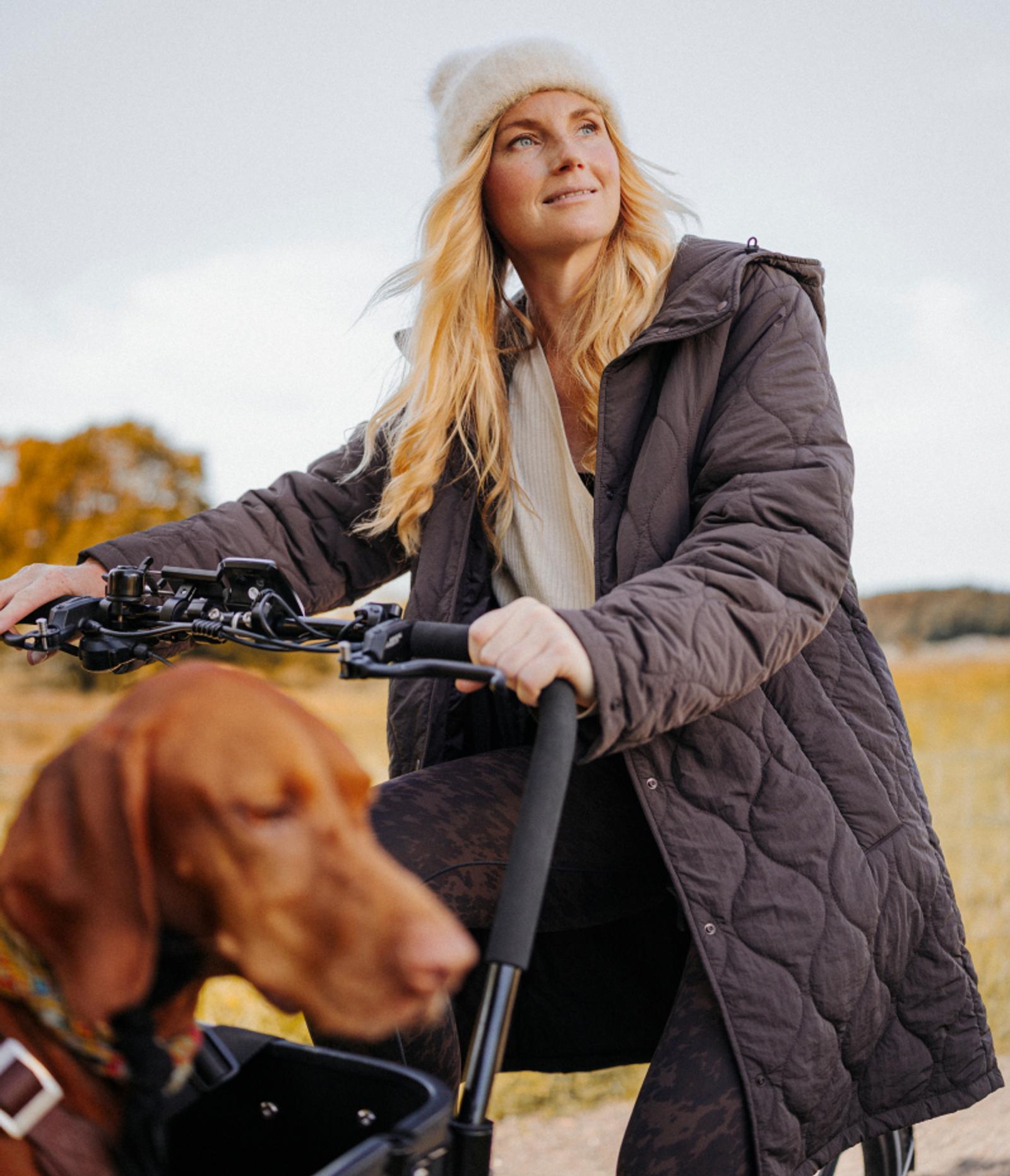 A woman on a bicycle with a dog in a front basket, looking to her right.