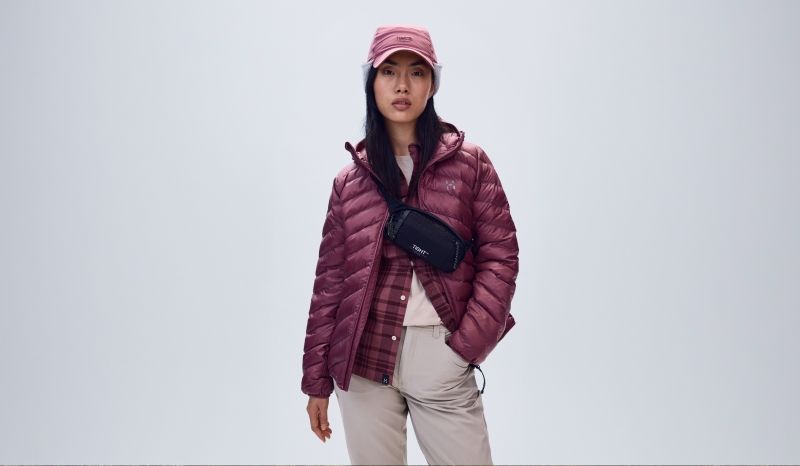 A person in a maroon puffer jacket, plaid shirt, beige cargo pants, pink baseball cap, and black crossbody fanny pack.