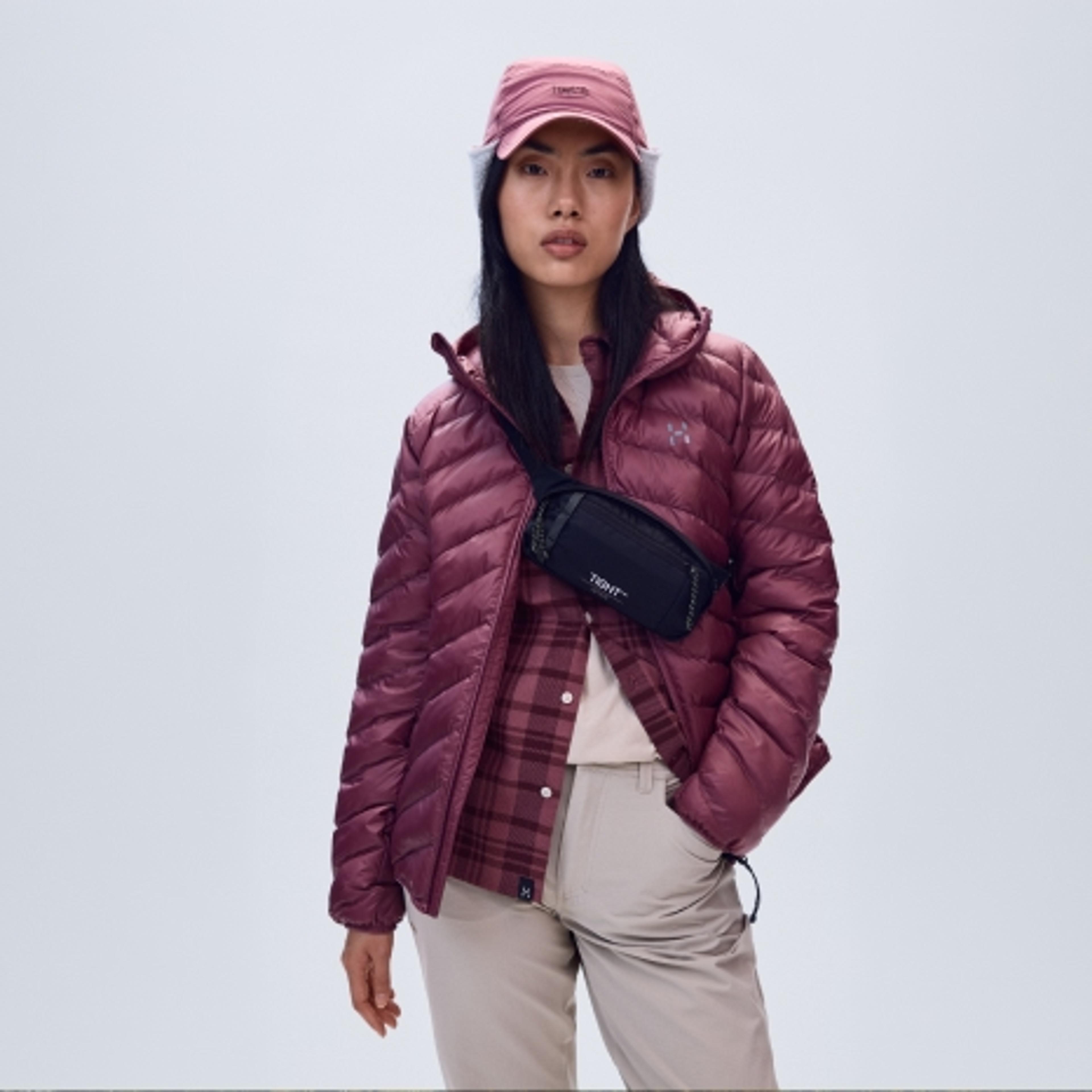 A person in a maroon puffer jacket, plaid shirt, beige cargo pants, pink baseball cap, and black crossbody fanny pack.