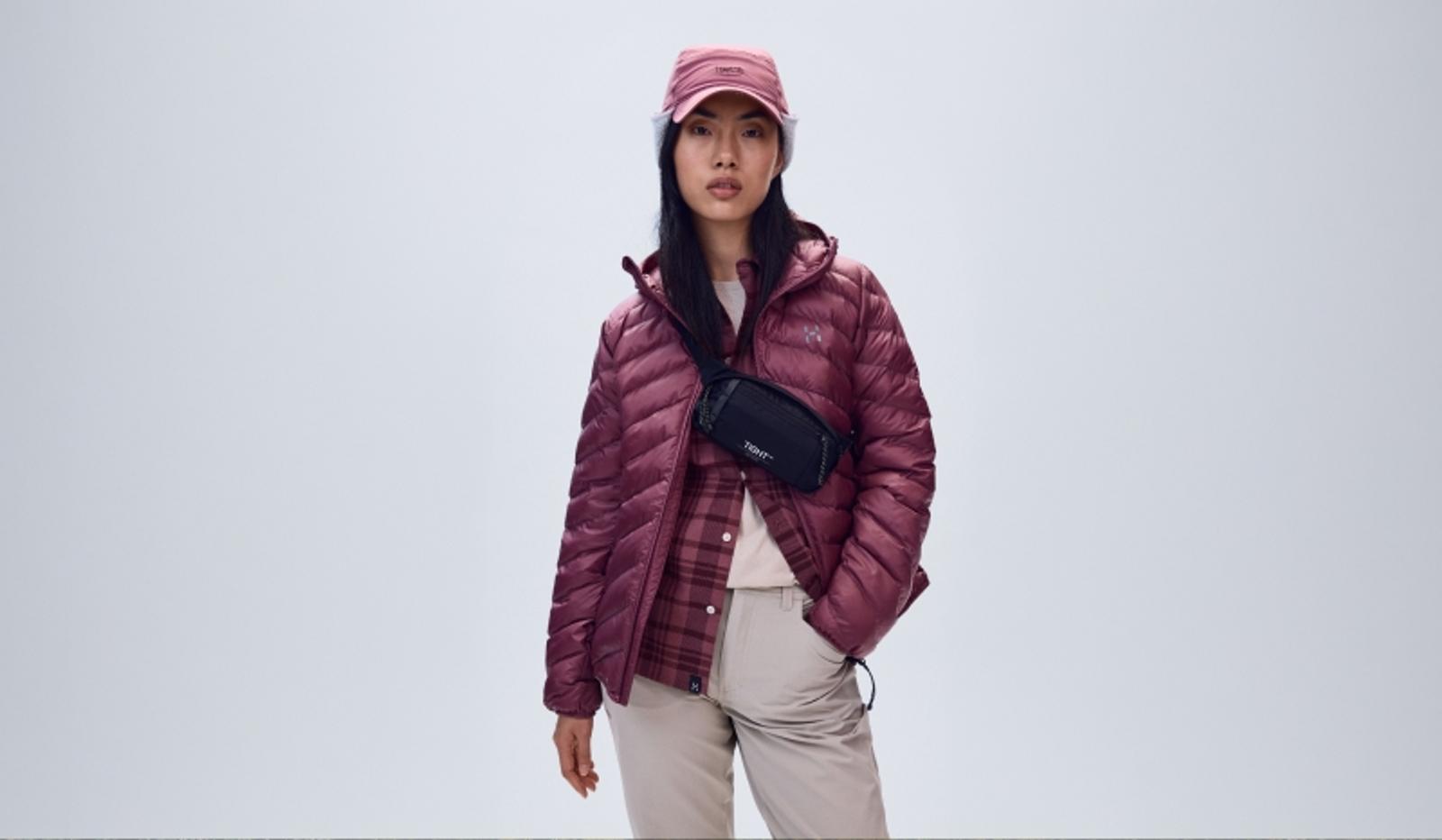 A woman wears a plum puffer jacket, pink plaid shirt, beige pants, pink cap, and black fanny pack.