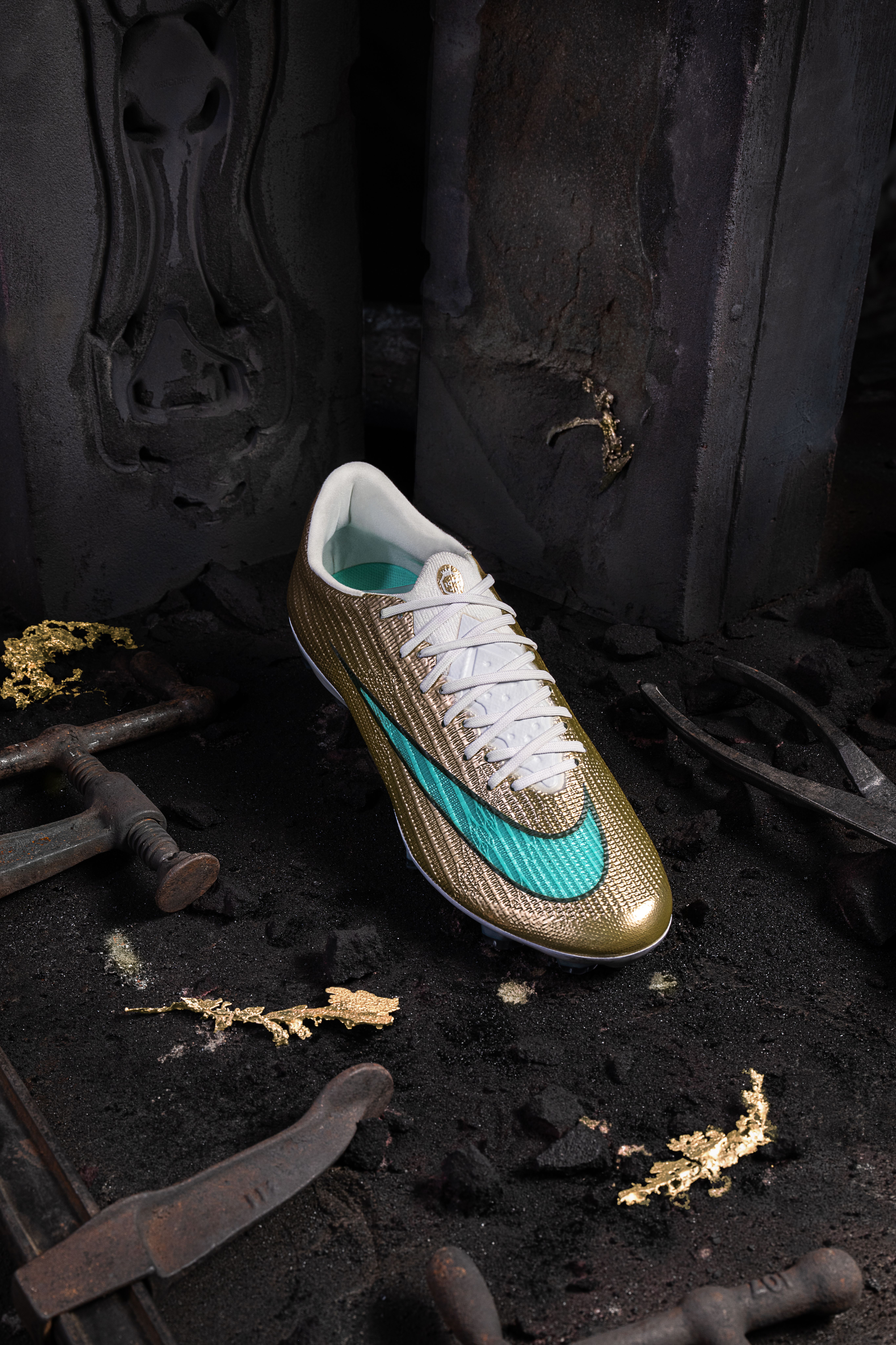 A gold soccer cleat with a teal Nike swoosh sits on dark ground with scattered gold flakes and old metal tools.