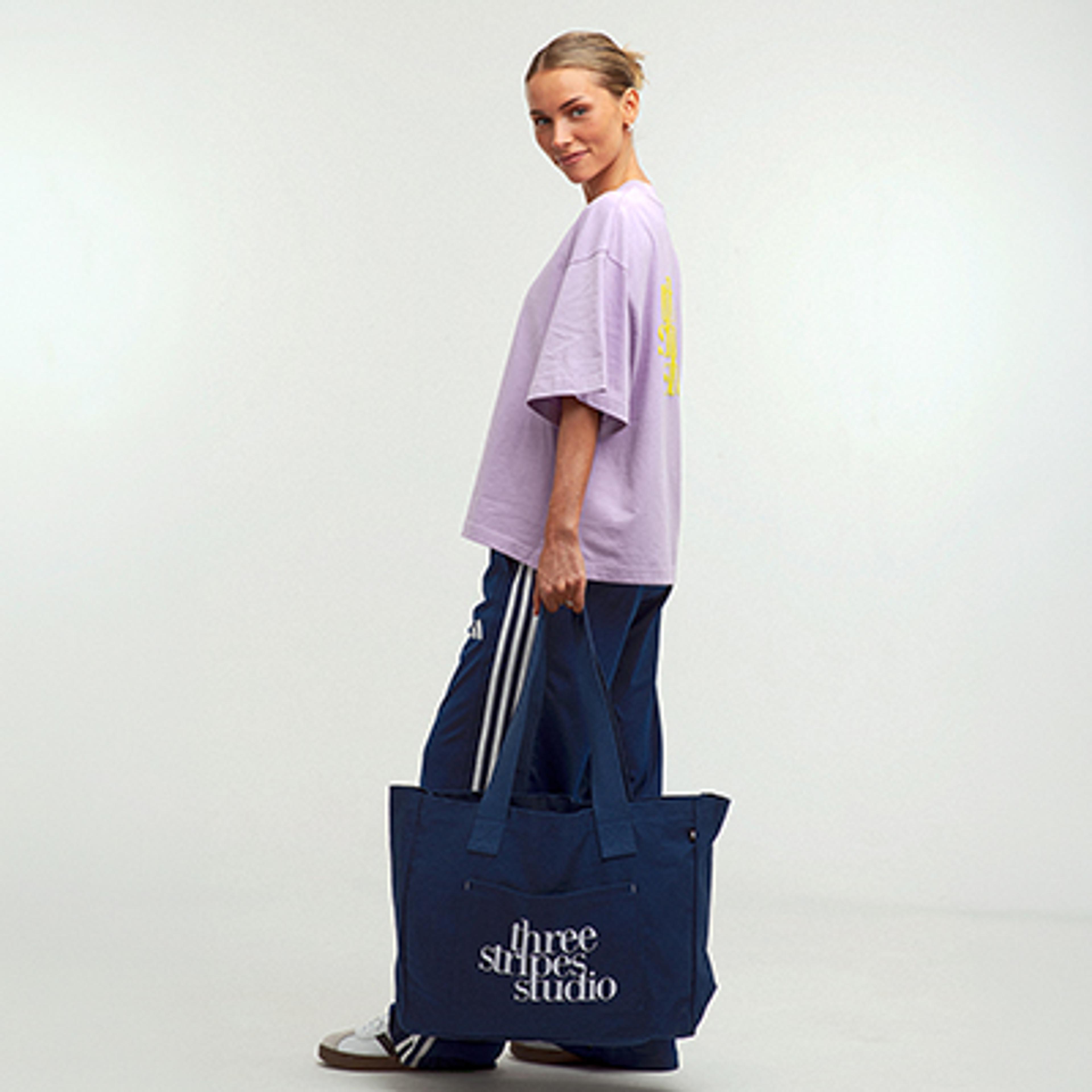 A woman in a purple oversized t-shirt and blue track pants holds a navy tote bag with "three stripes studio" text, smiling at the camera.