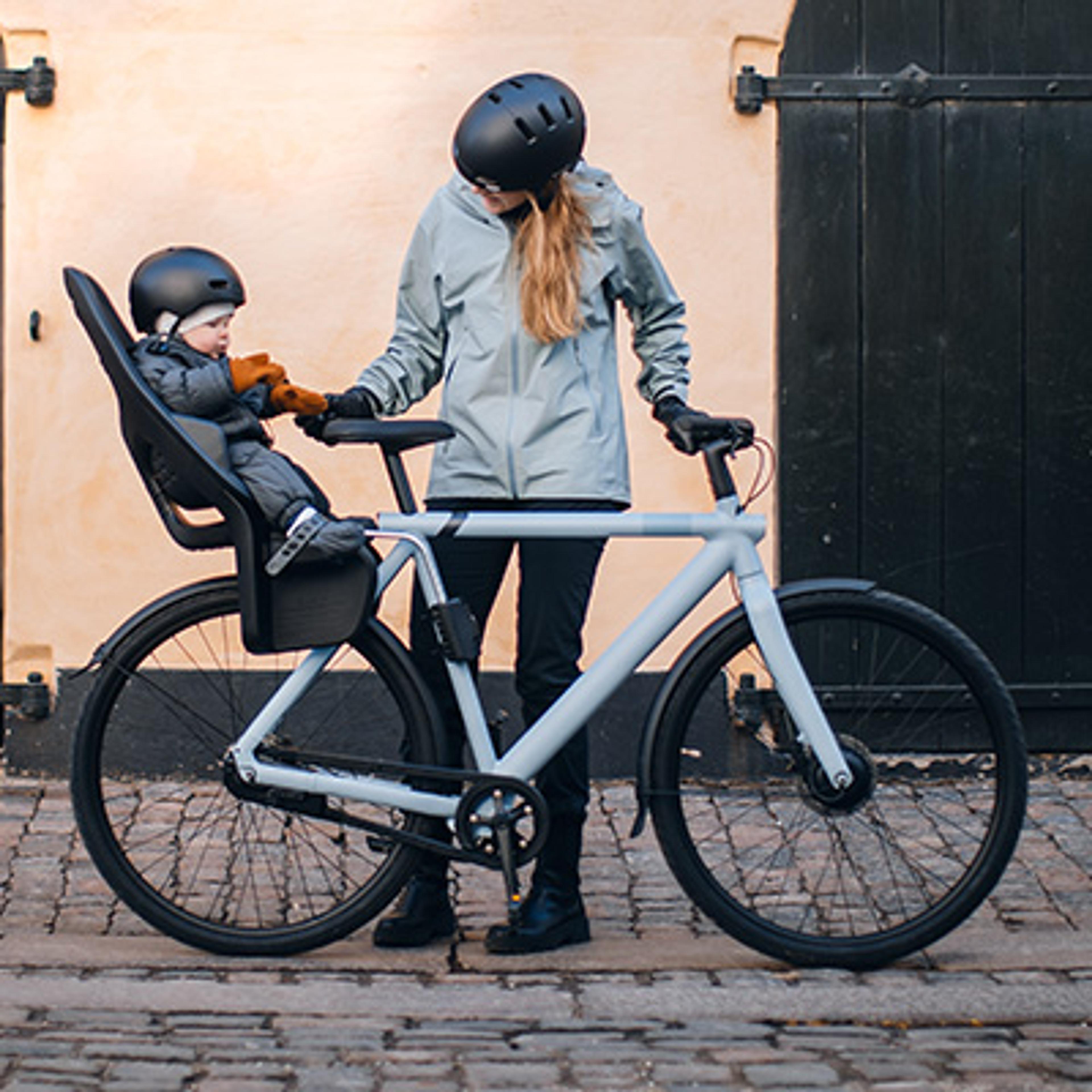 A woman and baby, both in helmets, next to a light blue bicycle with a child seat, holding hands.