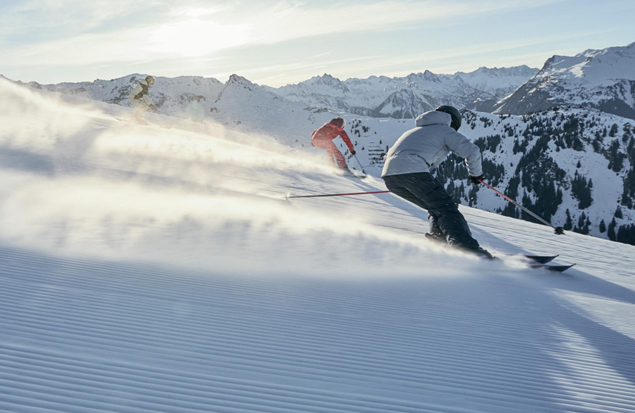 Three skiers carve down a sunny, groomed mountain slope, kicking up snow.