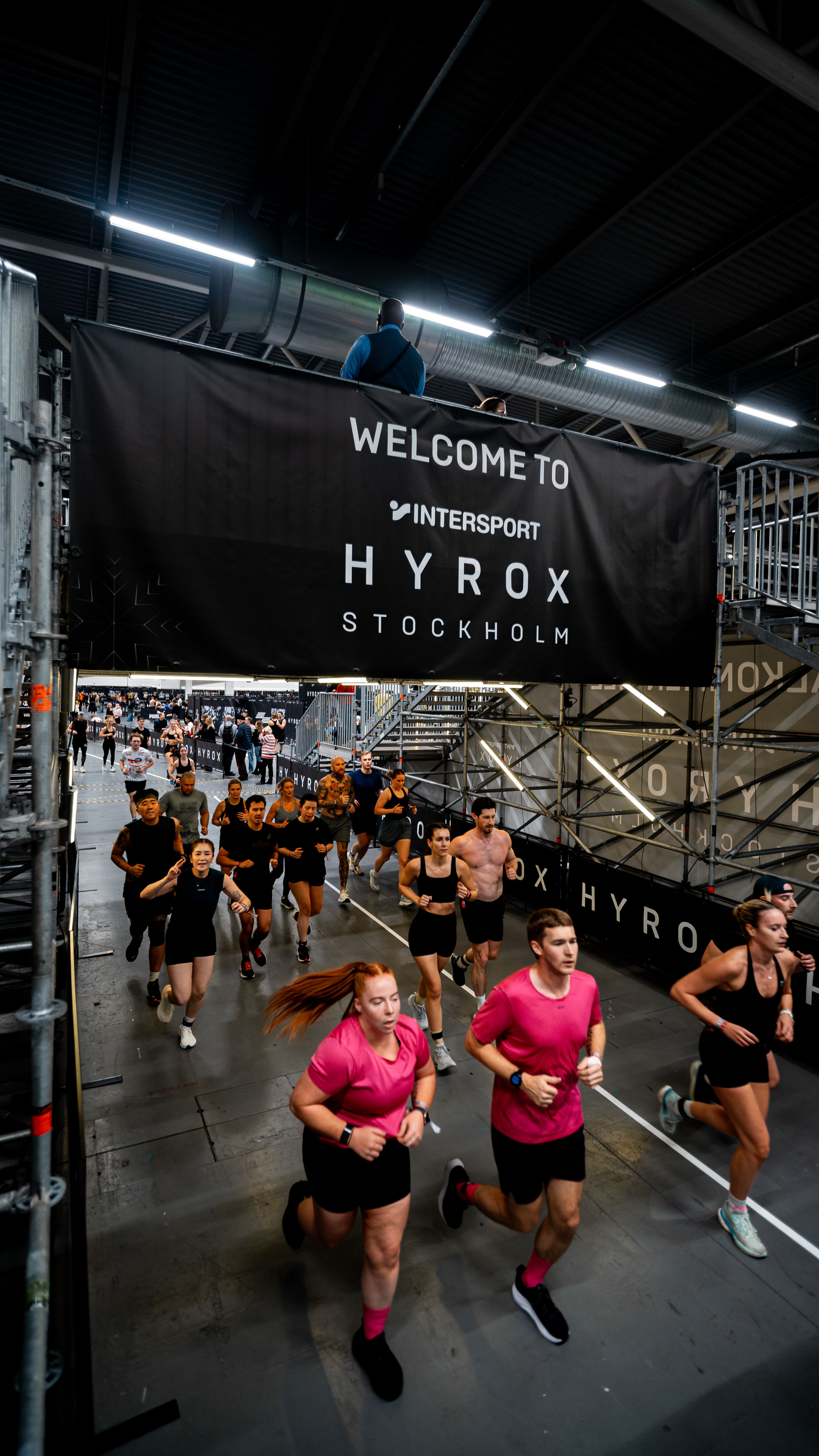 A group of people running indoors at a Hyrox Stockholm event, beneath a banner that reads "Welcome to Intersport Hyrox Stockholm".