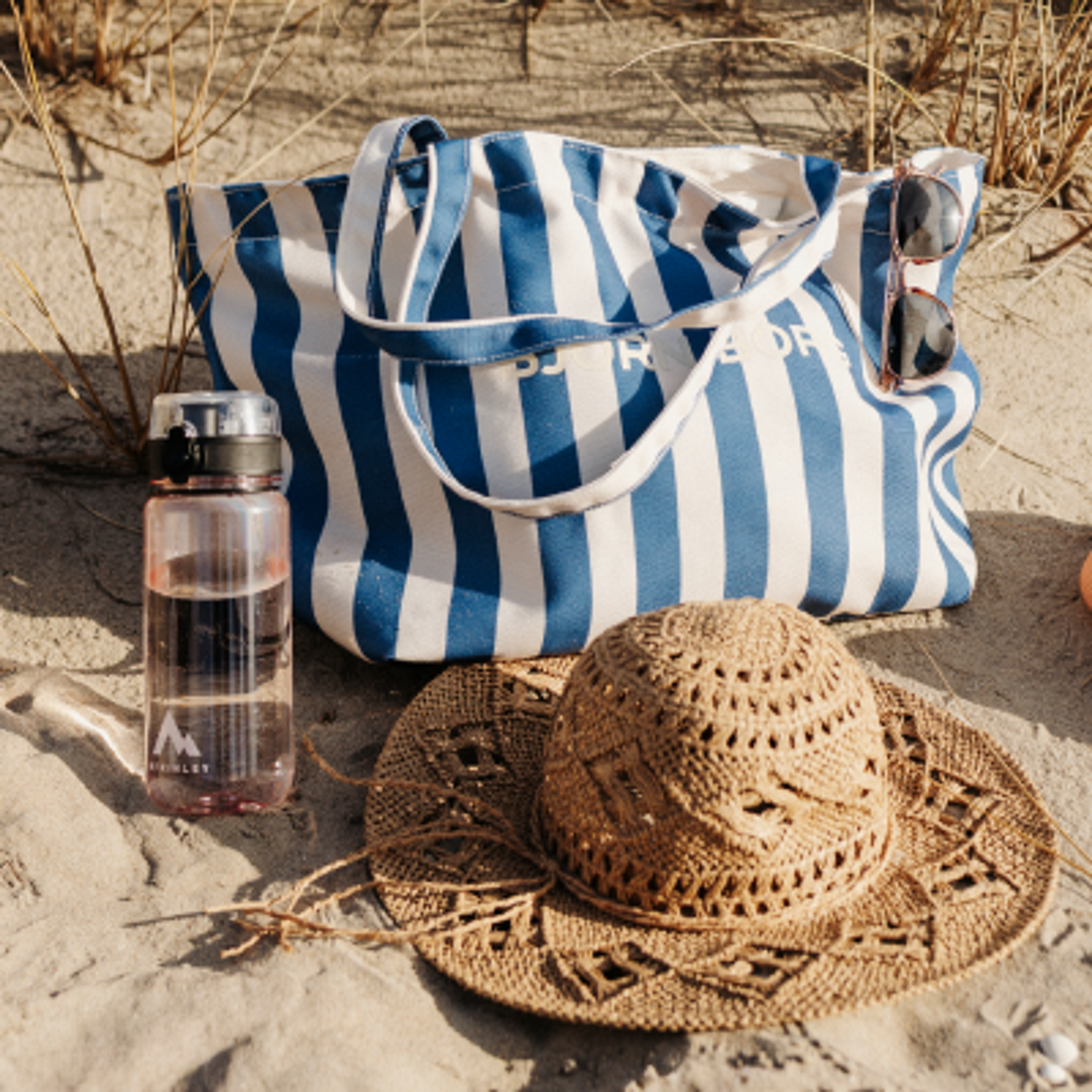 A blue and white striped beach bag, sunglasses, straw hat, and water bottle on sandy ground.