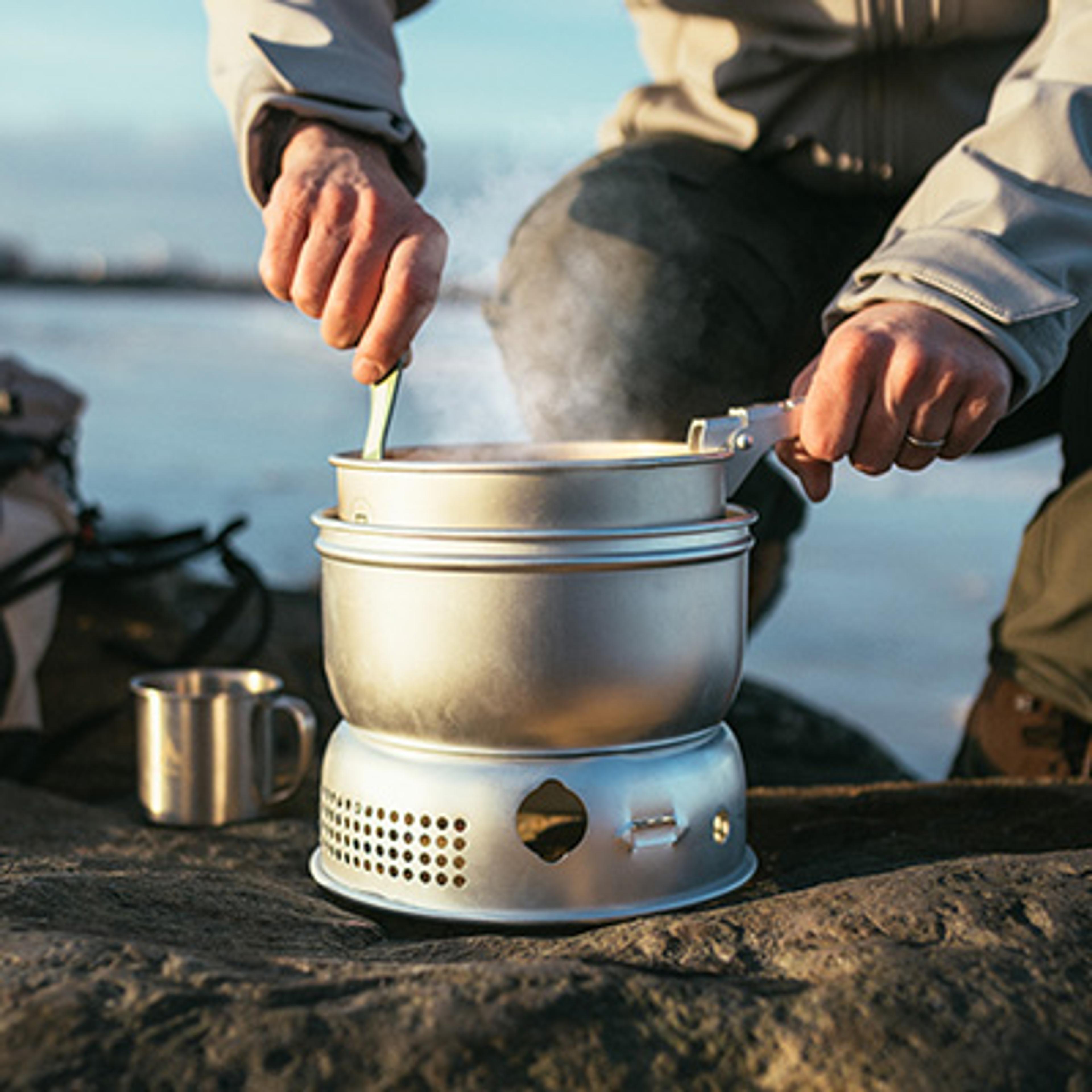 A person stirs steaming food in a portable camping pot on a stove outdoors, with a lake in the background.