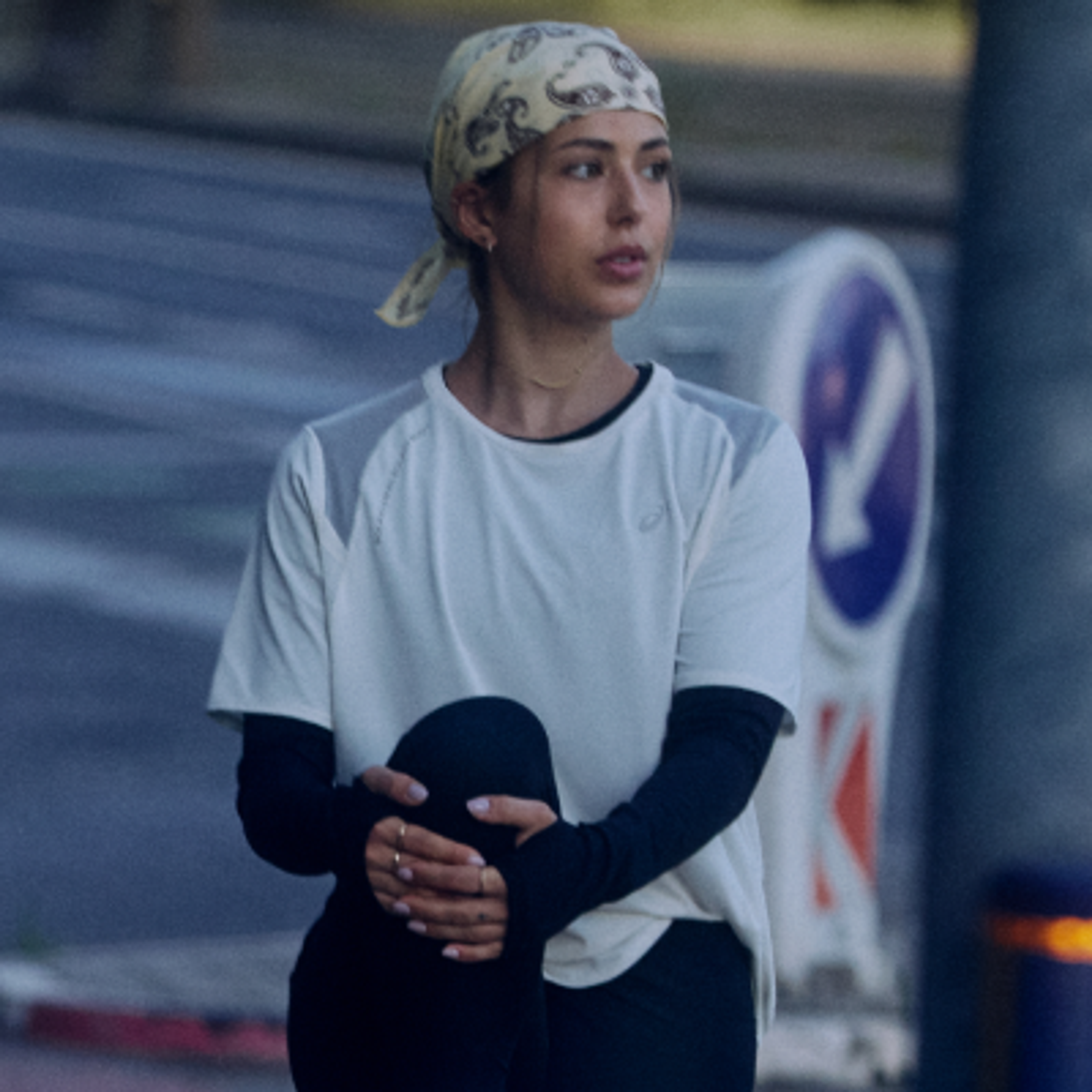 A female runner in a light purple ASICS shirt, sweating and looking down to catch her breath outdoors.