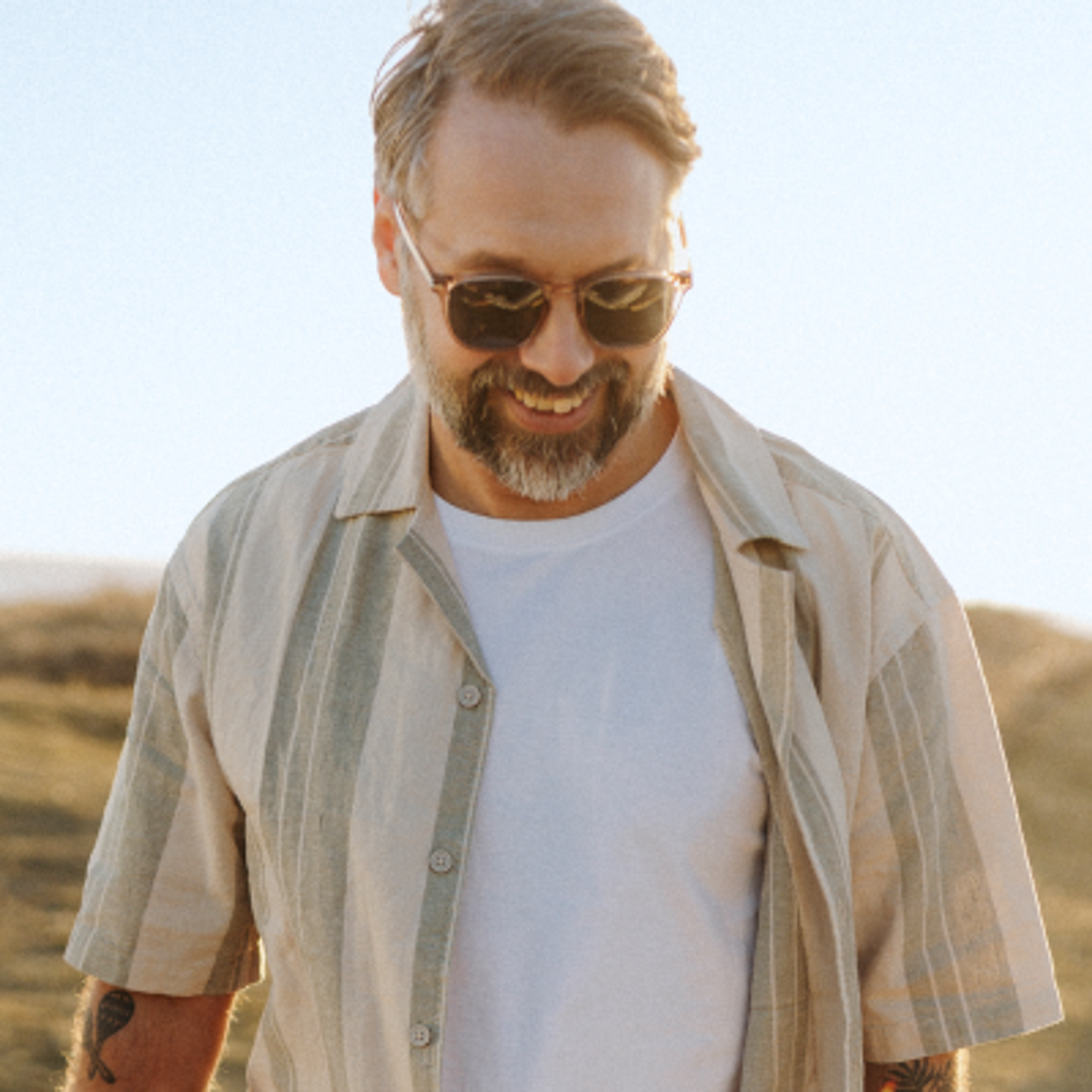 Smiling man with a beard and sunglasses, wearing a striped shirt, outdoors in golden light.