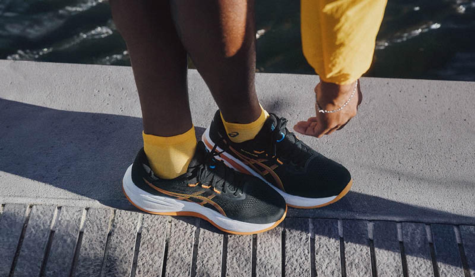 Person wearing black running shoes with orange details and yellow socks stands on concrete by water.