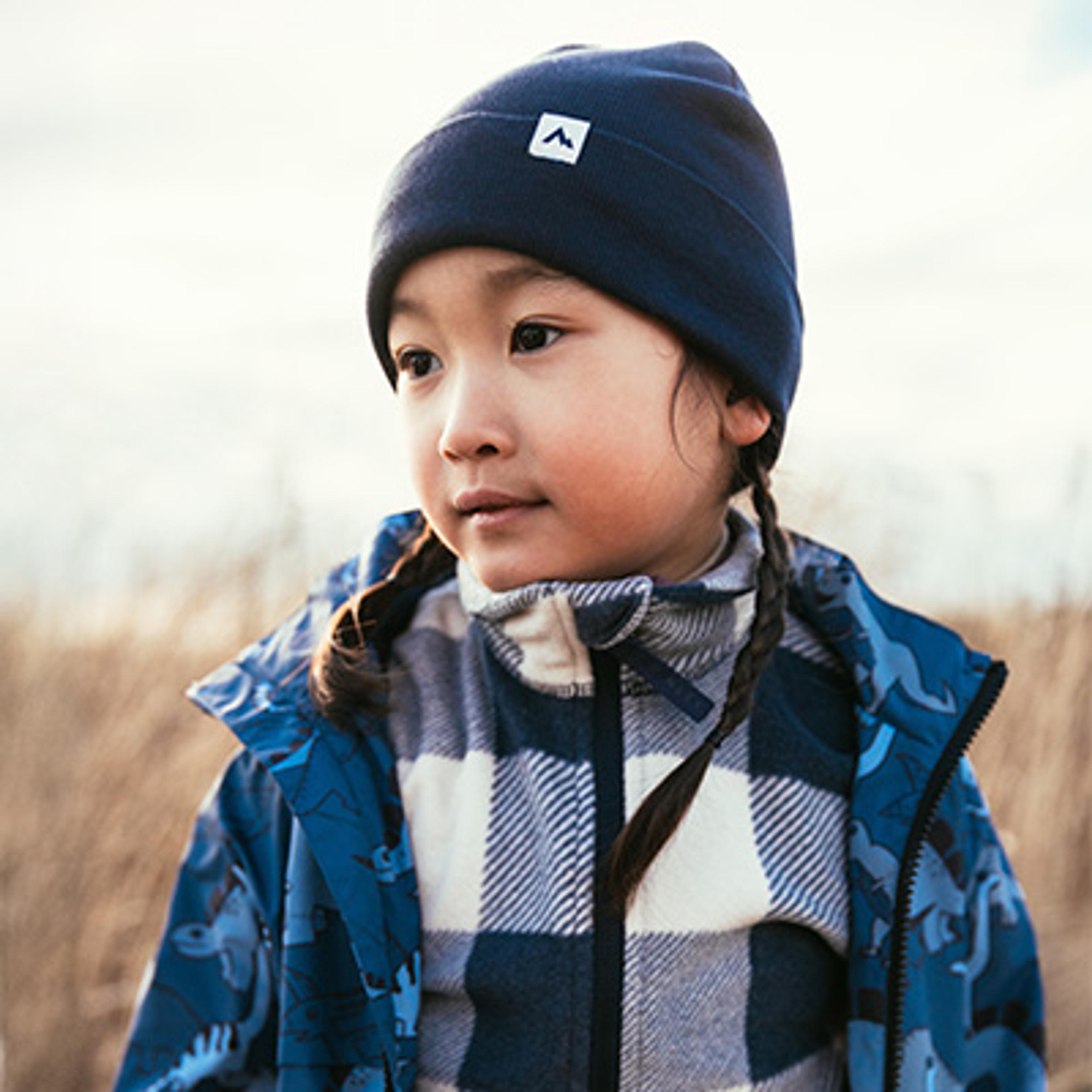 A young child with braids wears a blue beanie, plaid fleece, and a blue dinosaur-patterned jacket outdoors.
