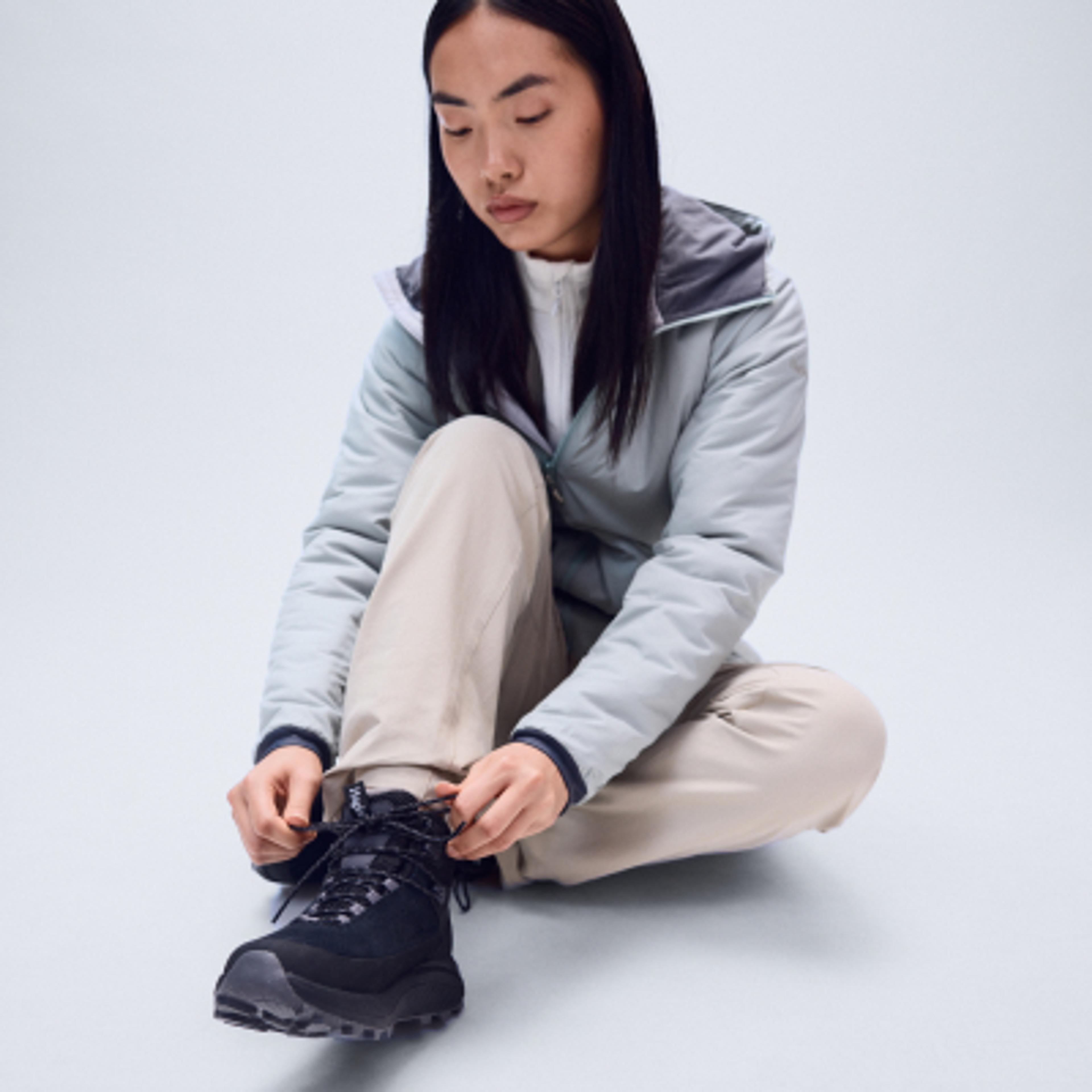 A person wearing a light blue jacket and tan pants sits on the floor, tying the laces of a black hiking boot.