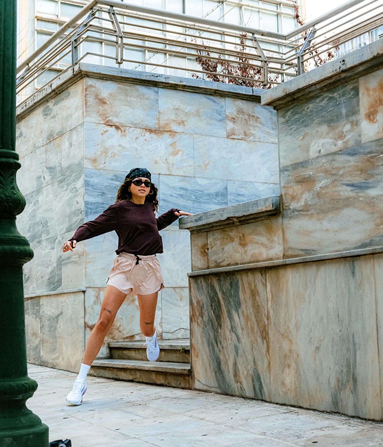 A woman in a bandana, sunglasses, dark top, and pink shorts leaps with arms outstretched against marble walls.