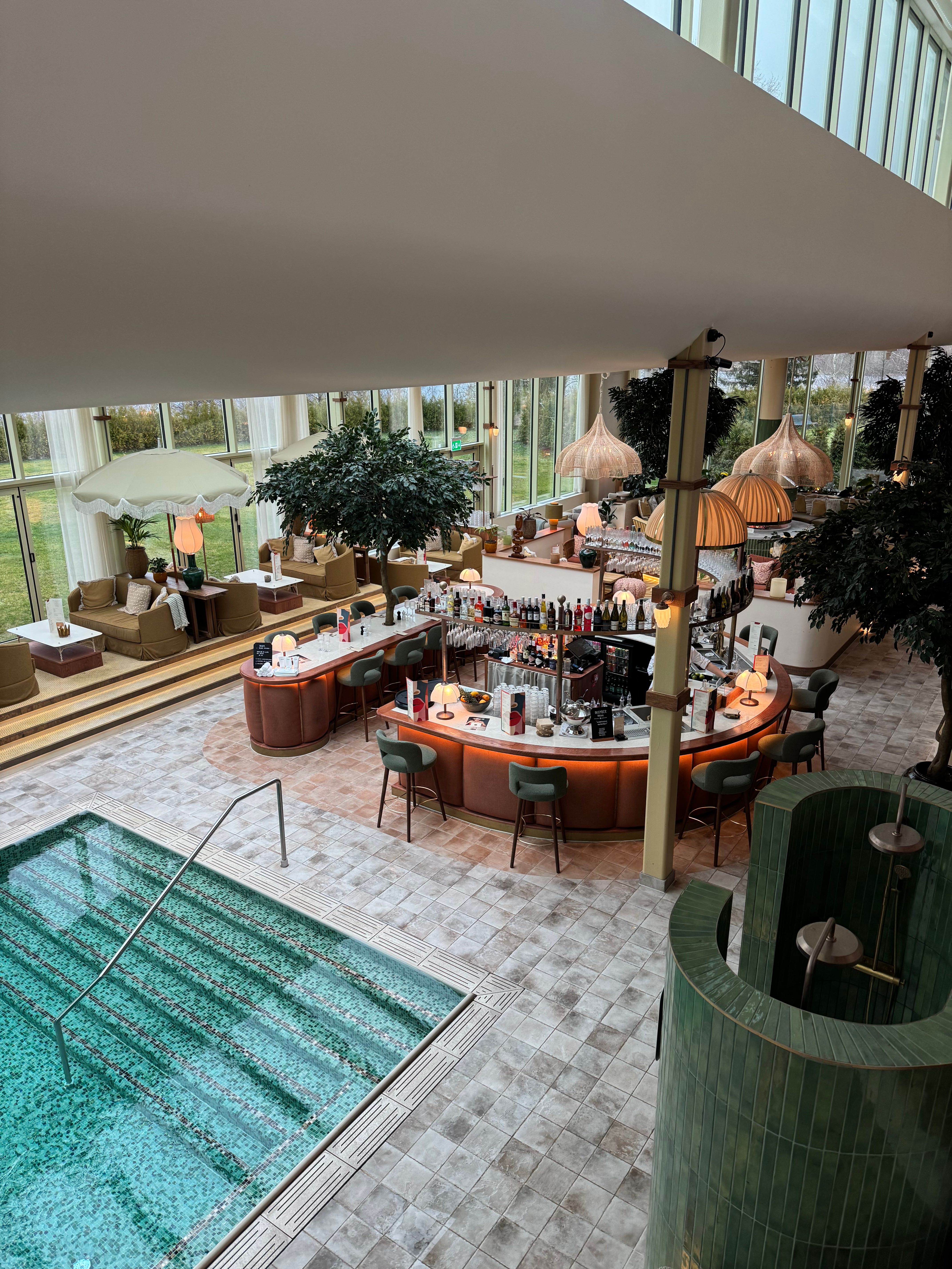 View from above of a large, stylish indoor space with a turquoise-tiled swimming pool, a circular bar, and lounge seating by tall windows.