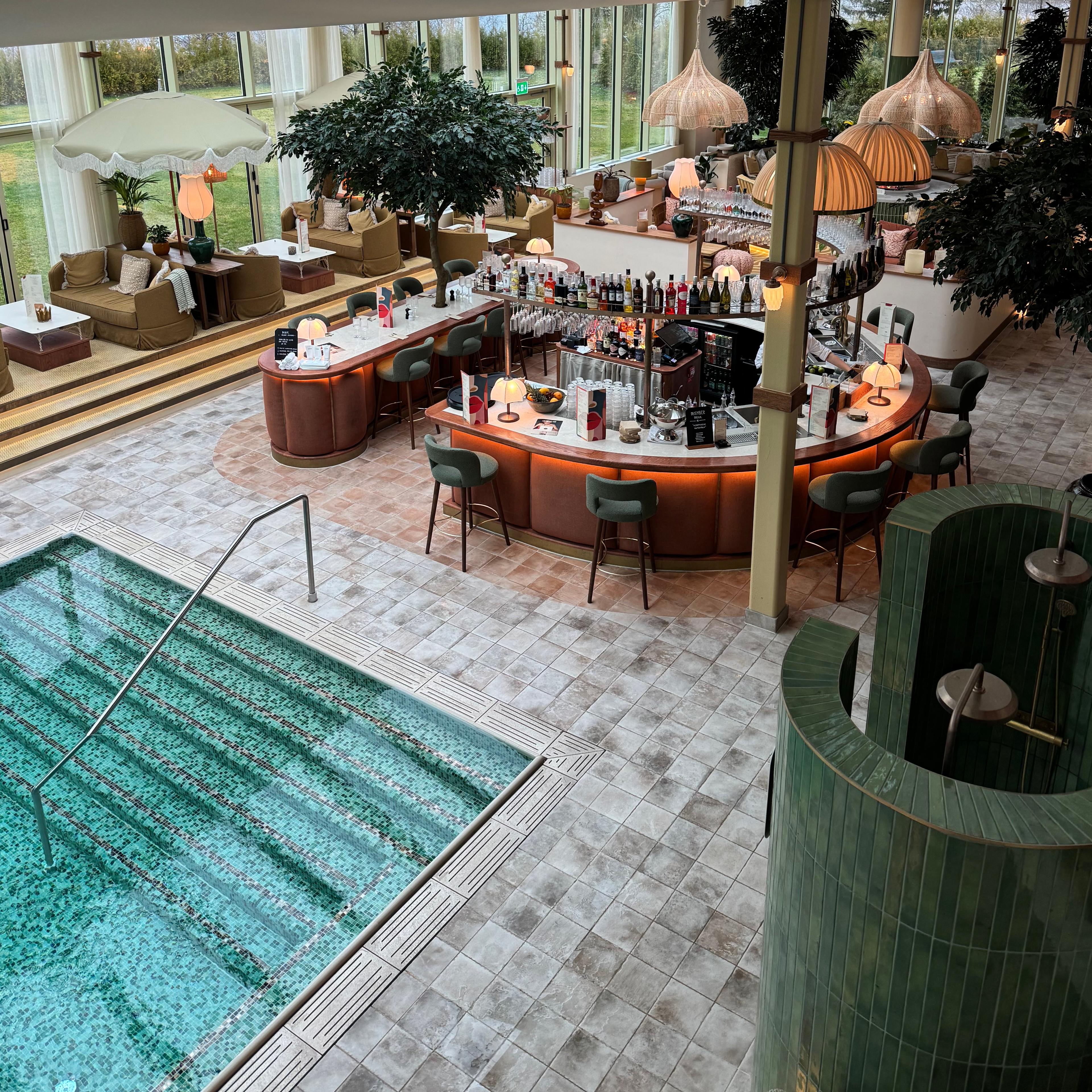 View from above of a large, stylish indoor space with a turquoise-tiled swimming pool, a circular bar, and lounge seating by tall windows.