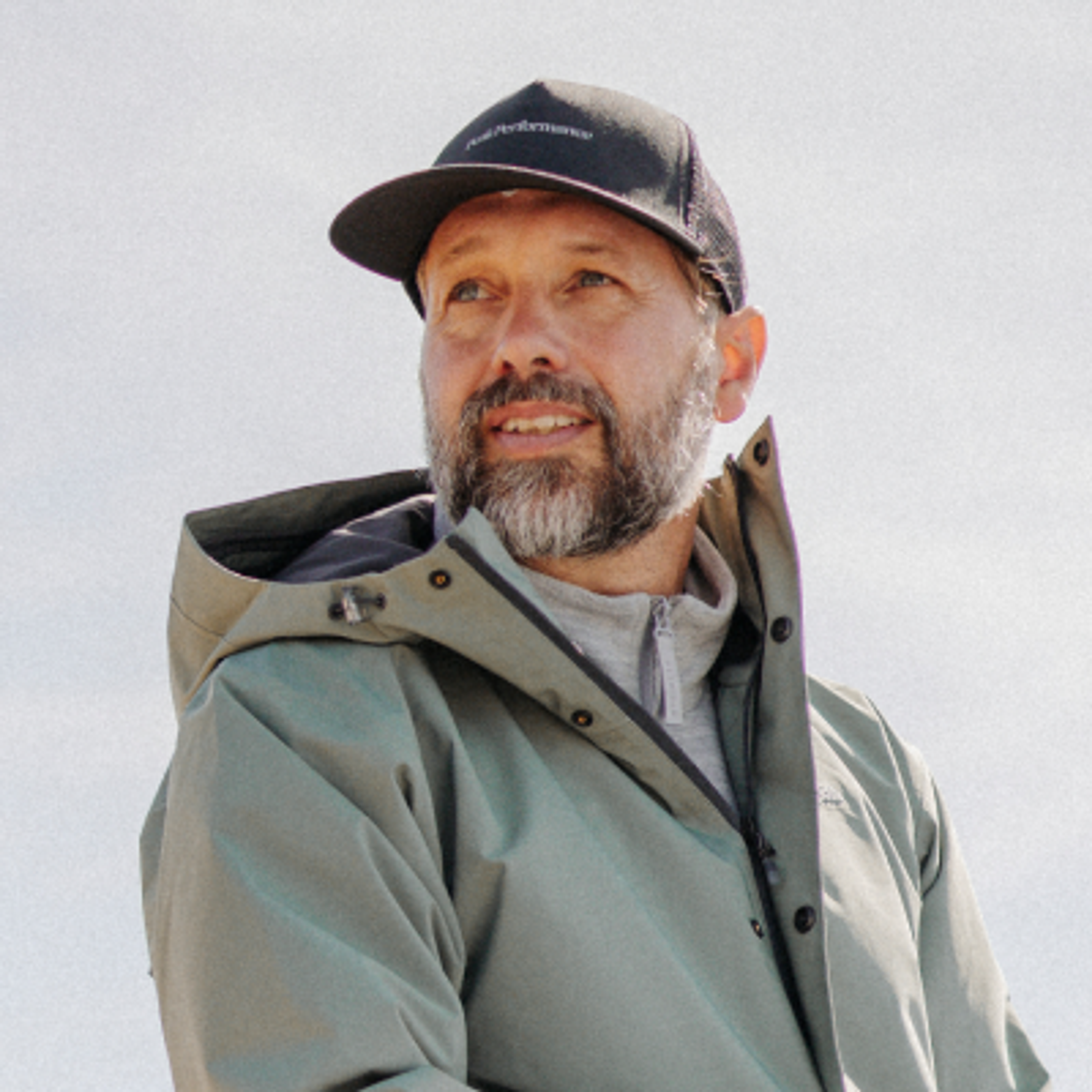 A smiling man with a beard wears a black "Peak Performance" cap and a light green jacket, looking upward.