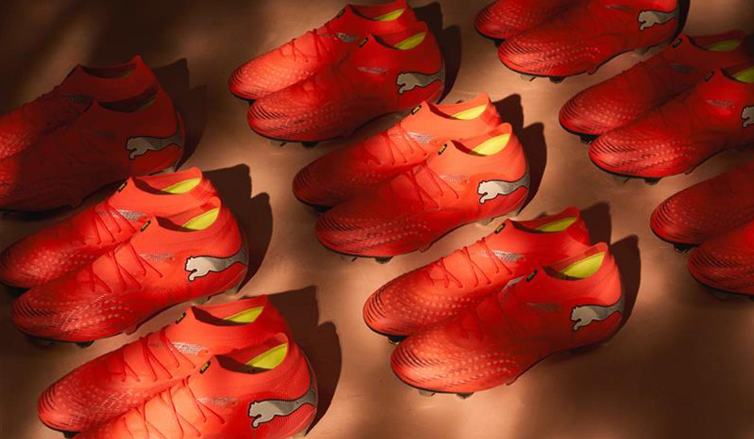 Rows of red Puma soccer cleats with silver logos and green trim on a brown surface with shadows.