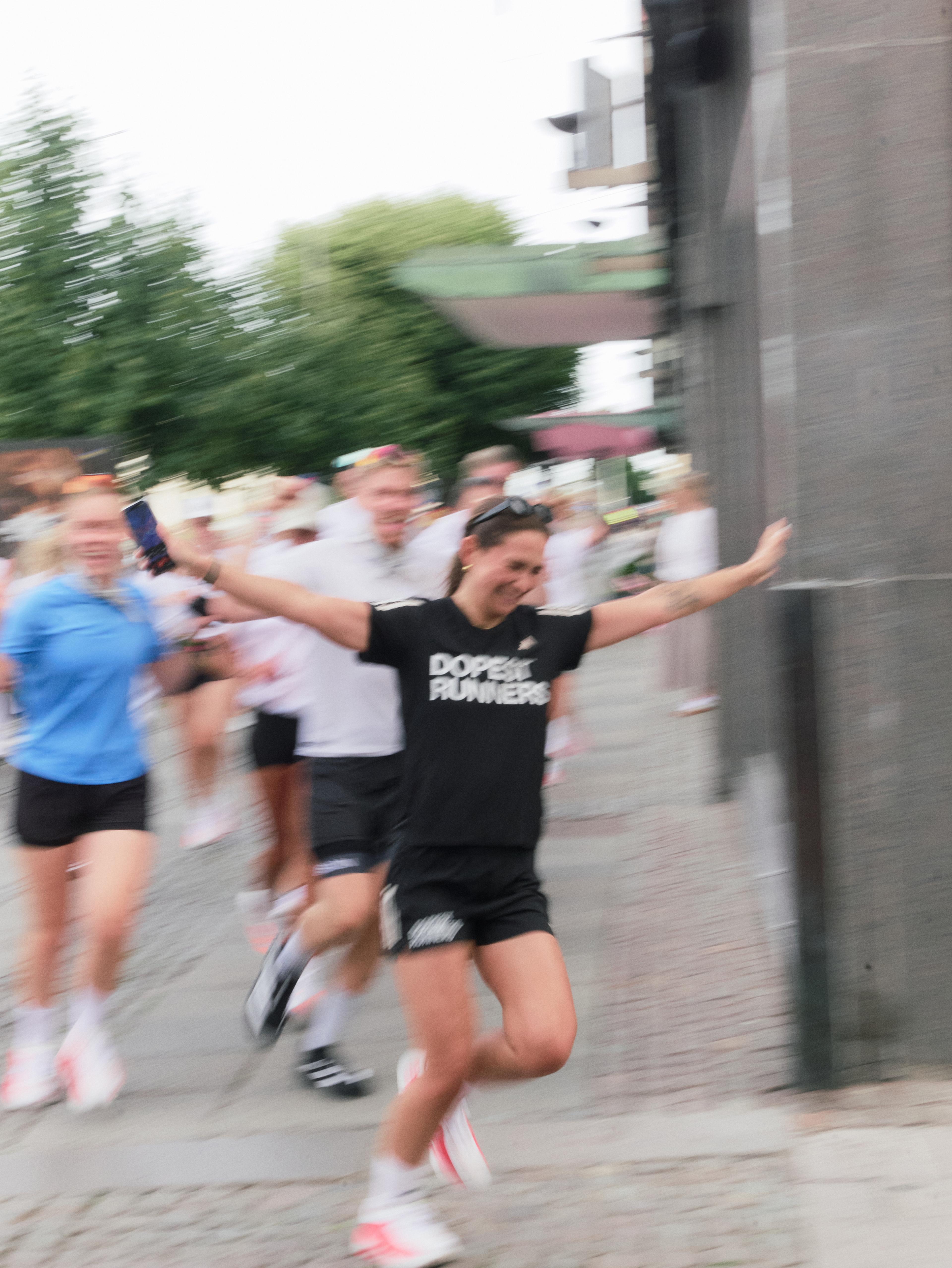 A smiling woman in a "DOPE RUNNERS" t-shirt runs with outstretched arms, captured with motion blur among other runners.