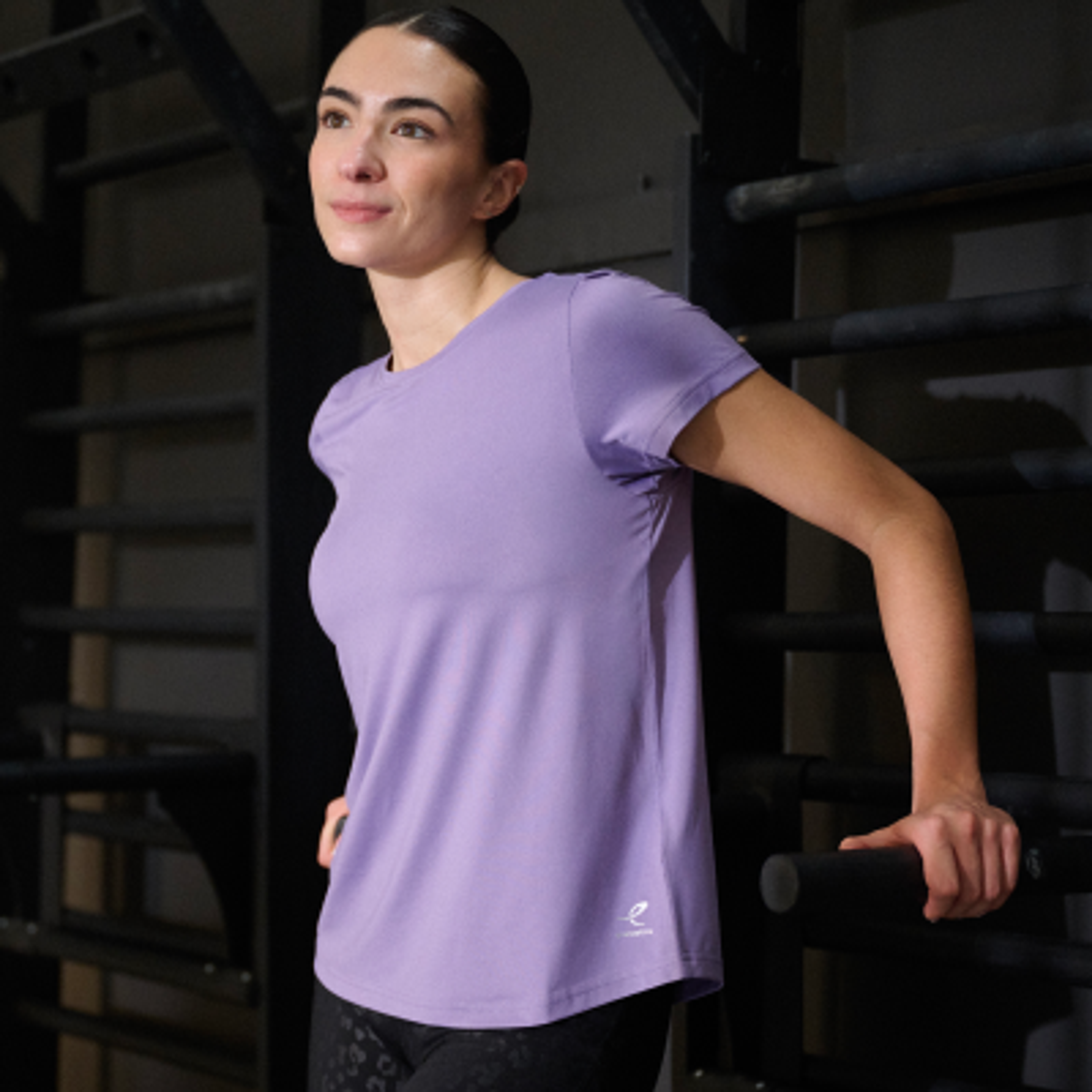 A woman in a light purple t-shirt and dark patterned leggings stands next to a gym rack, holding a bar.