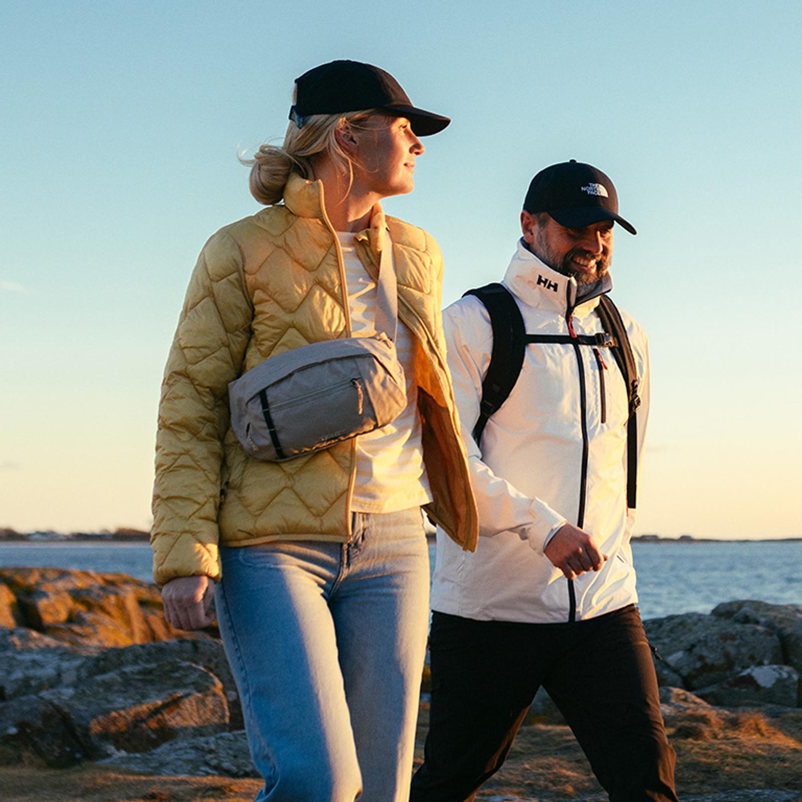 A woman in a yellow jacket and a man in a white jacket walk along a rocky coast at sunset.