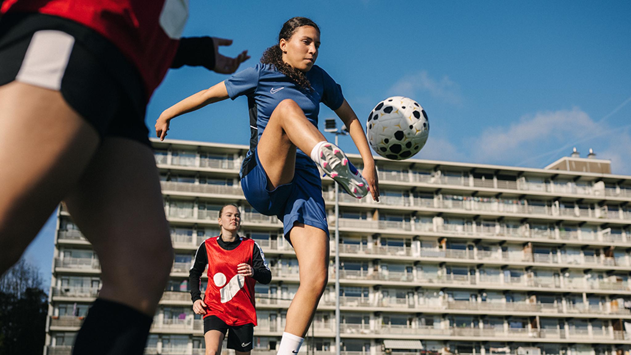 A female soccer player in a blue uniform juggles a black and white ball on an outdoor field.