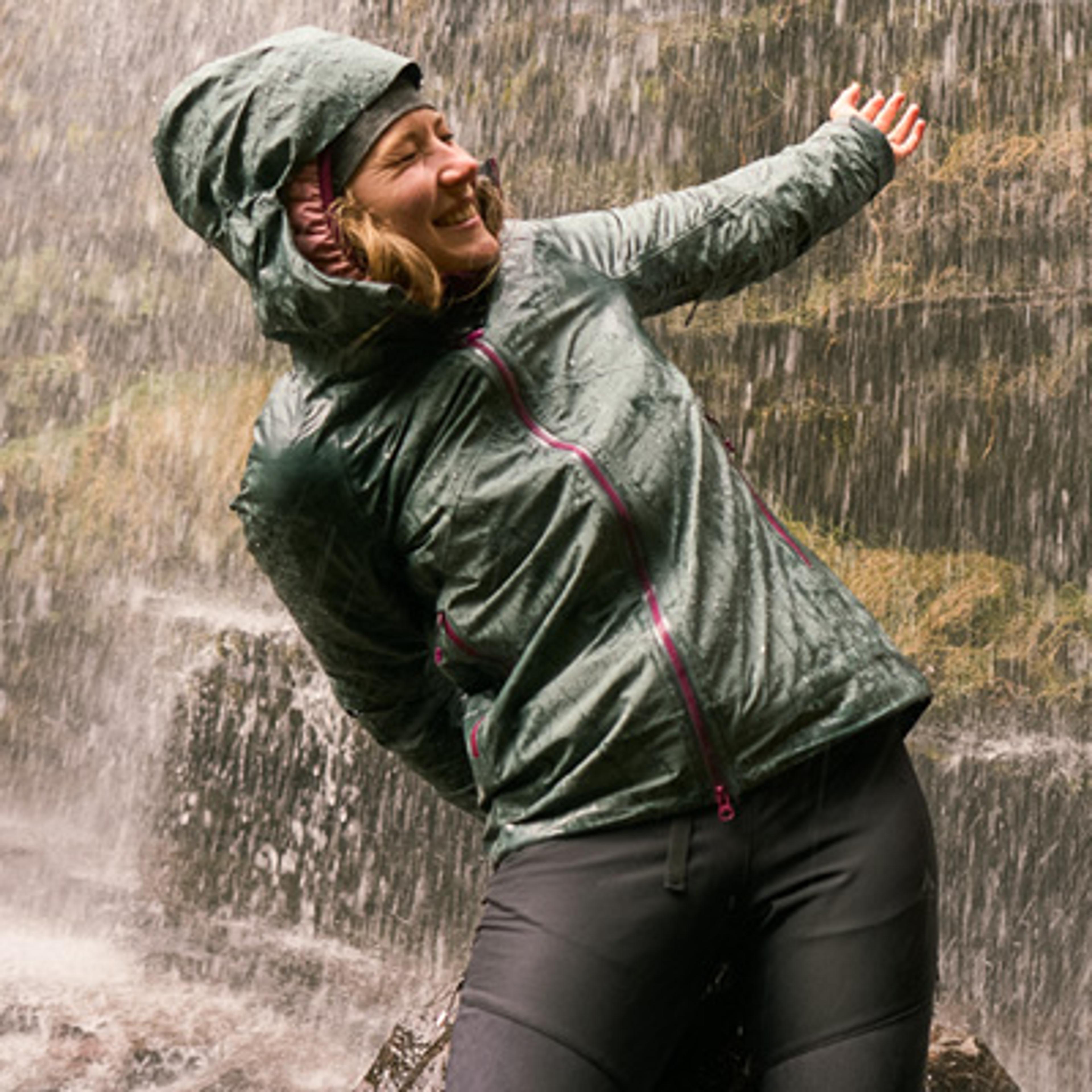 A smiling woman in a green waterproof jacket stands under a waterfall with one arm outstretched.