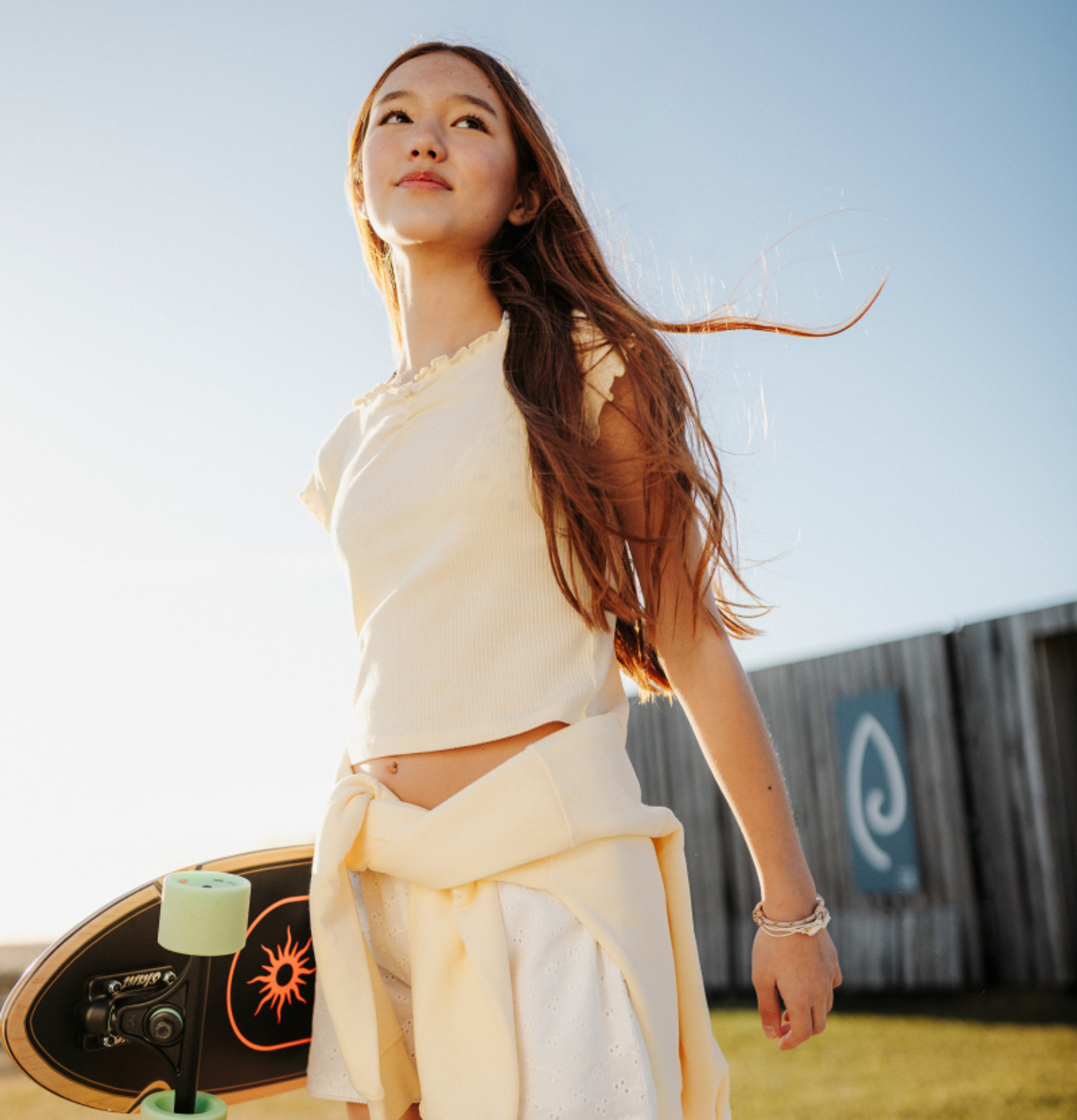 A young woman with long hair looks up while holding a skateboard on a sunny day.