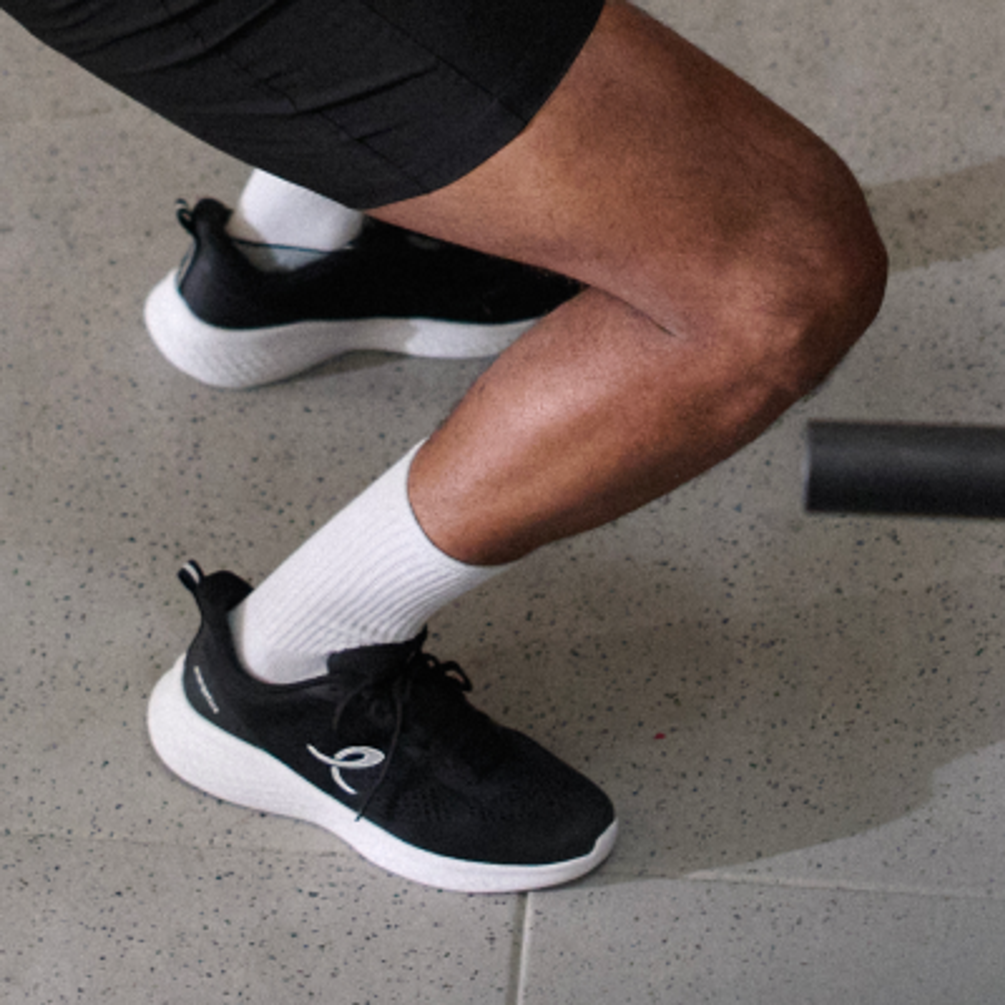 A person's legs in black shorts, white socks, and black sneakers with white soles, bent at the knees on a grey floor.
