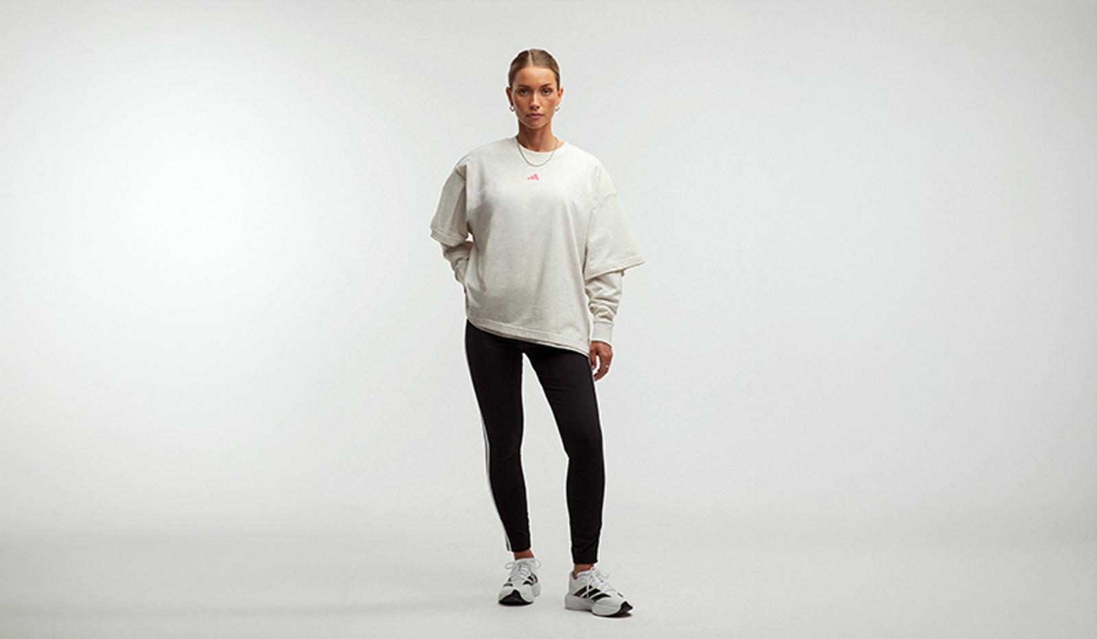A woman wears an oversized light grey Adidas sweatshirt, black striped leggings, and white sneakers against a white background.