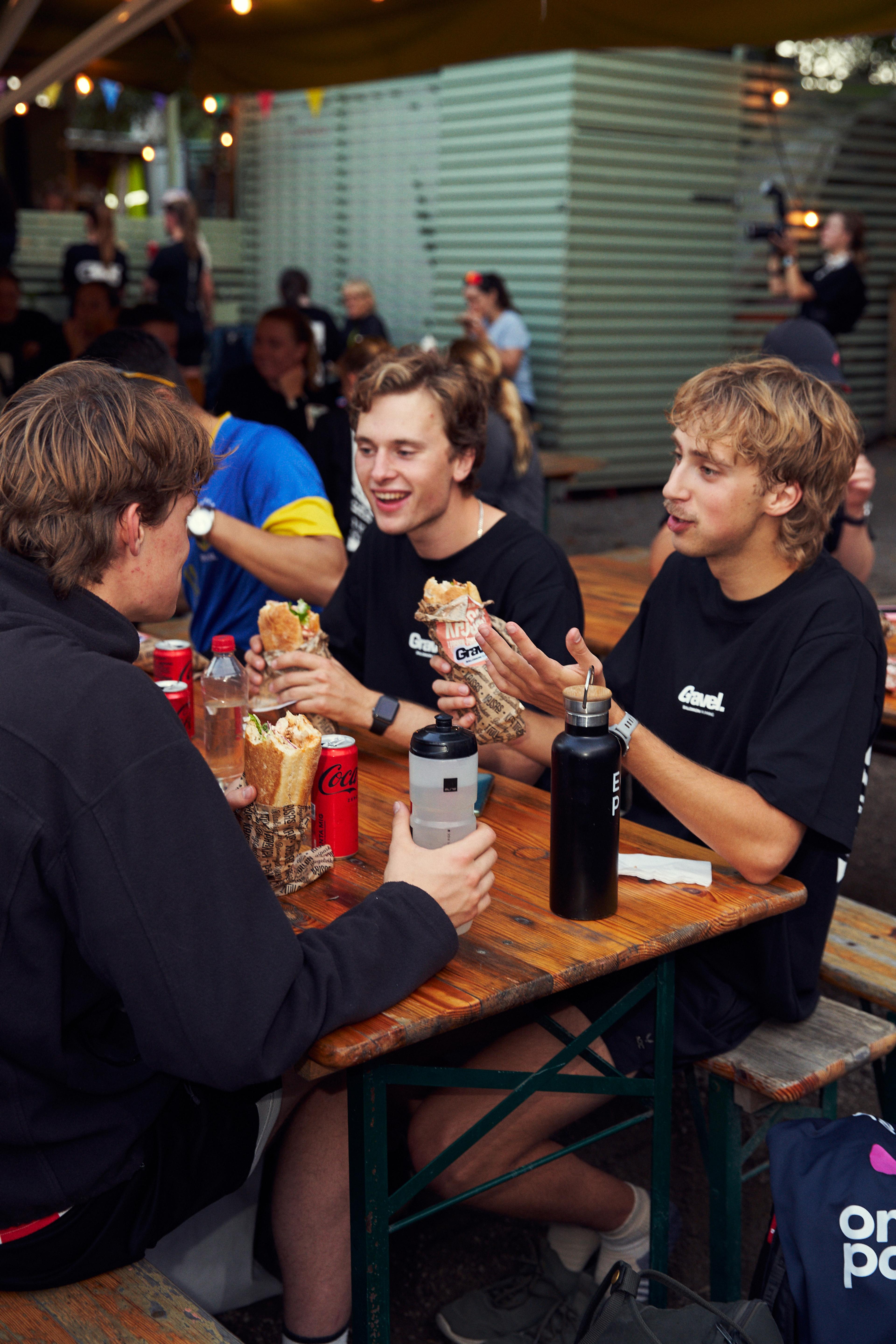 three young men are sitting at a table eating sandwiches and drinking coca cola