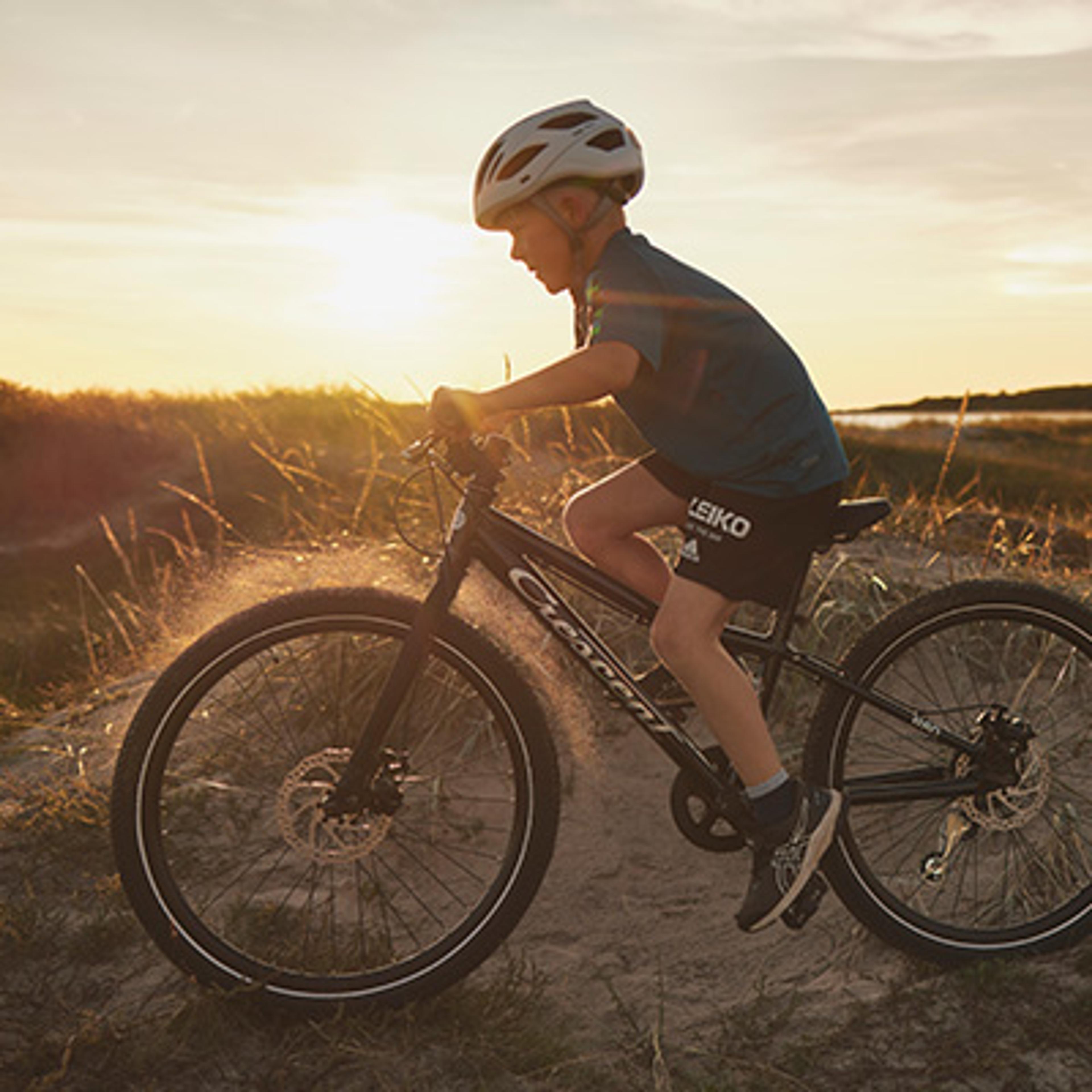 Boy in helmet riding a bicycle on a sandy path at sunset, kicking up dust.