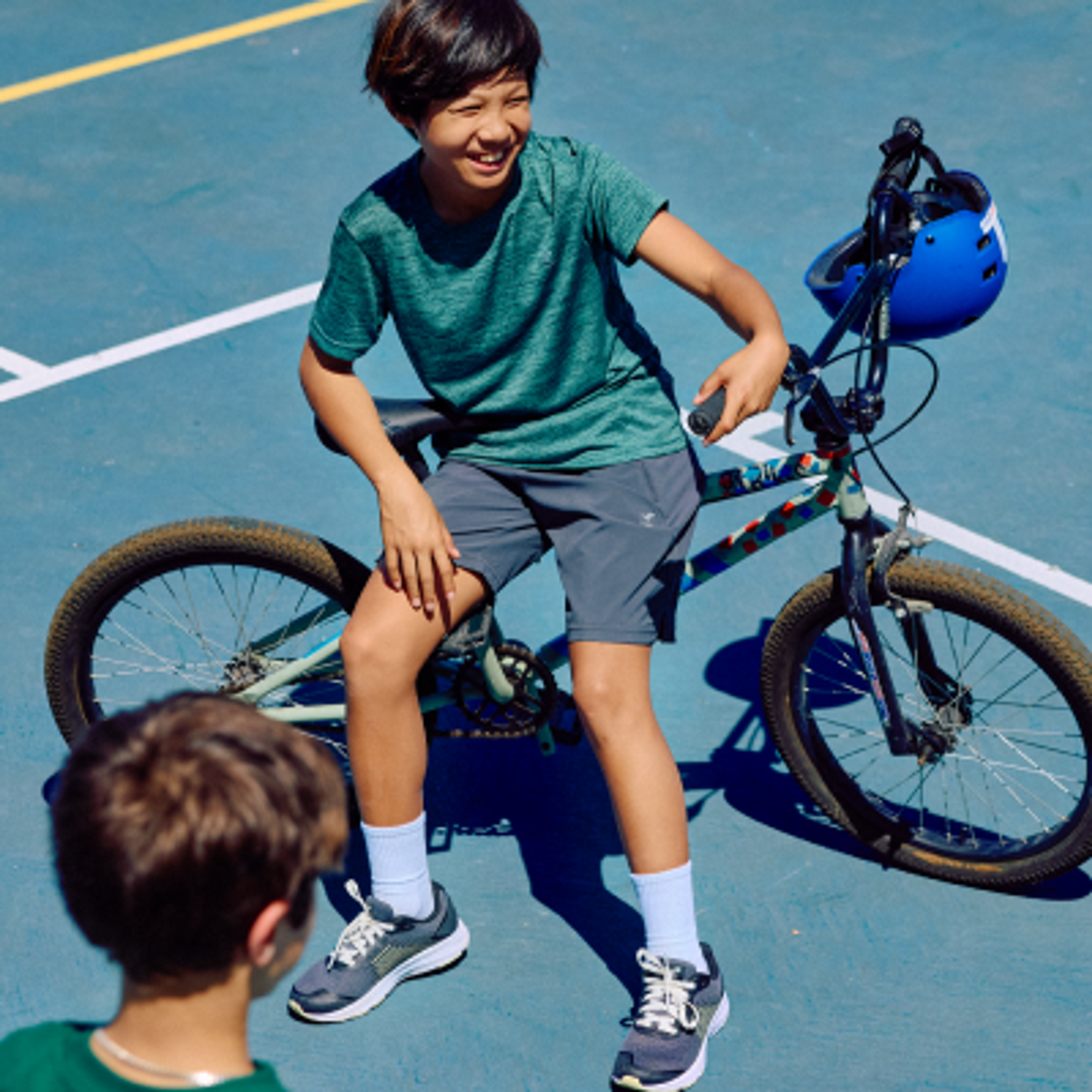 A smiling boy sits on a BMX bike with a blue helmet on the handlebars, facing another boy on a blue court.