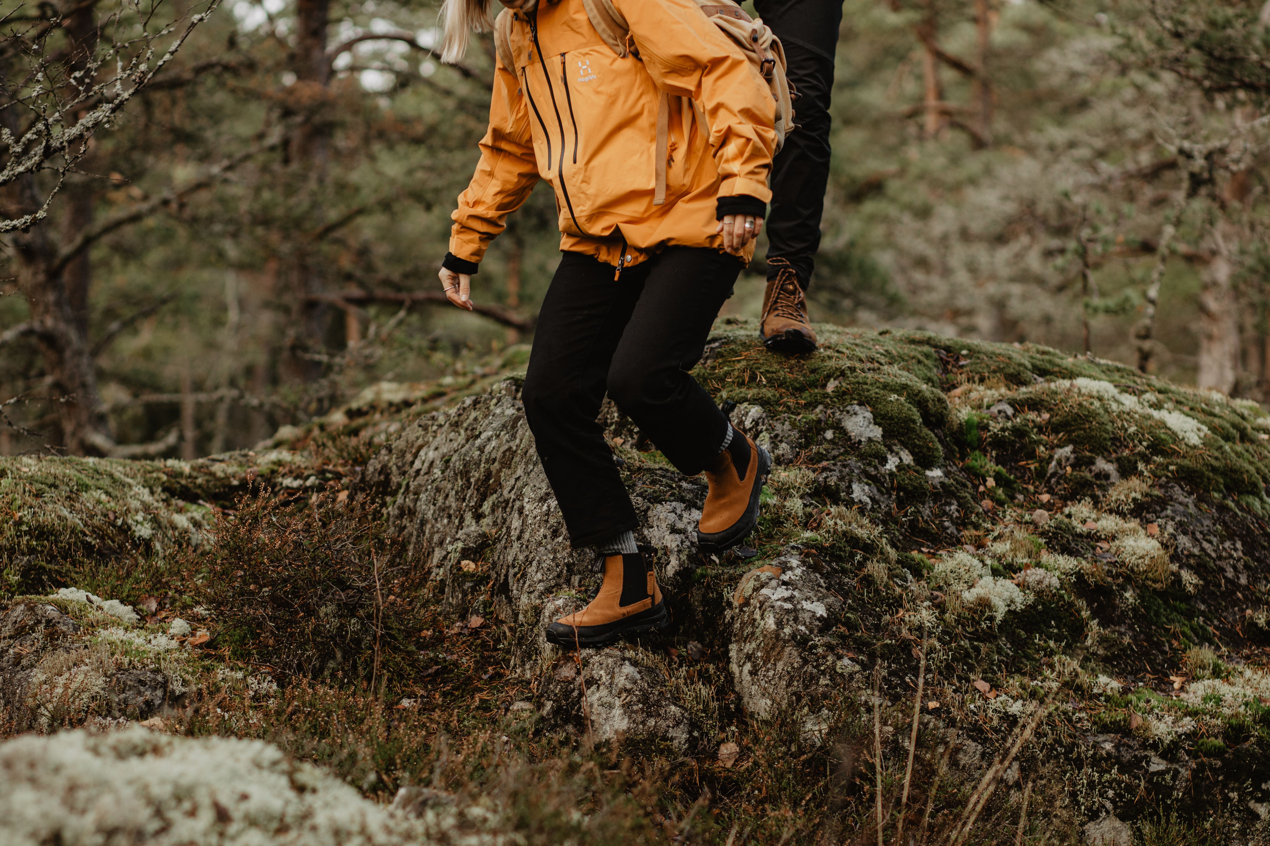 Two hikers, one in a yellow jacket and brown boots, descend a mossy rock in a forest.