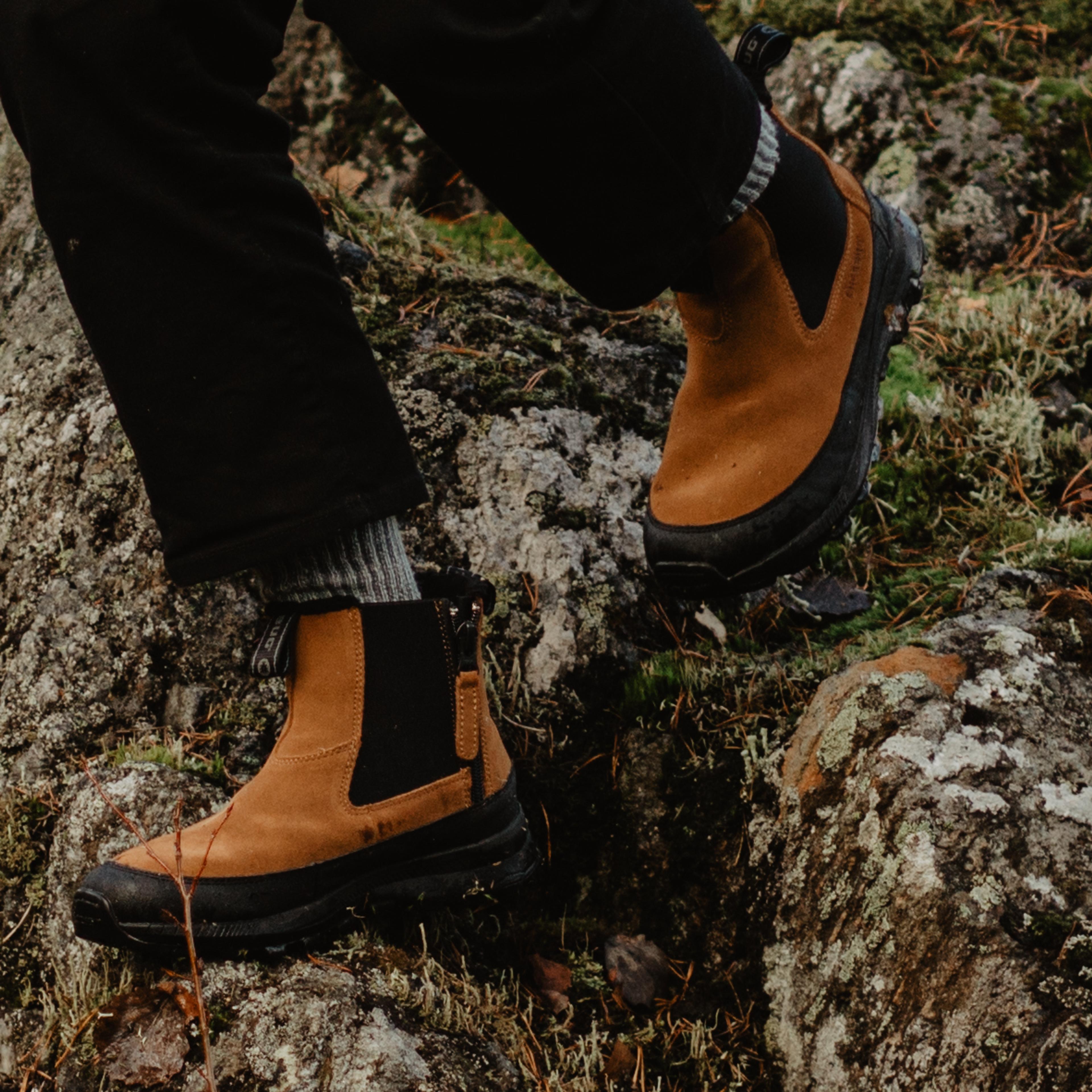 Two hikers, one in a yellow jacket and brown boots, descend a mossy rock in a forest.