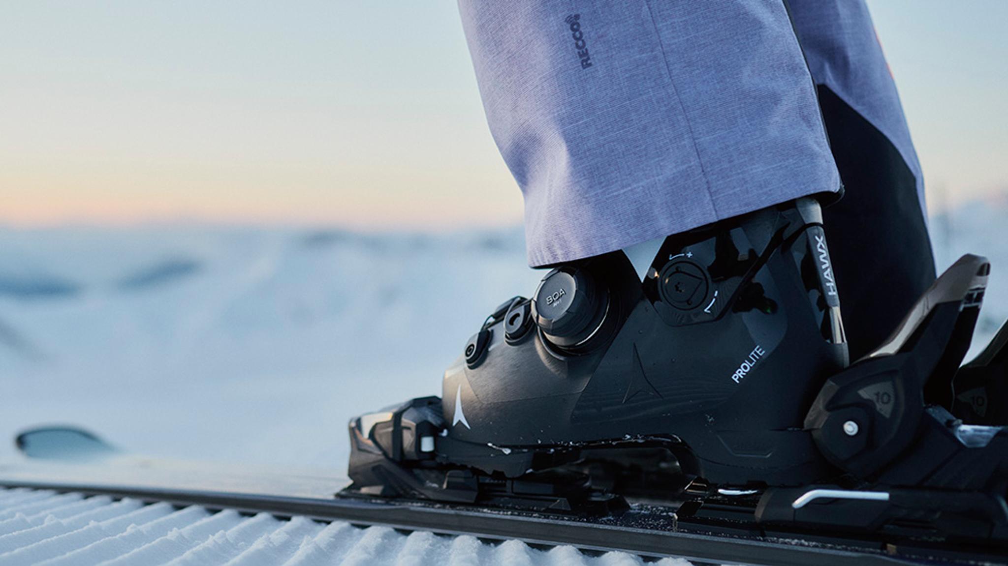 A black Atomic HAWX PROLITE ski boot on a ski, on groomed snow with a person's leg and snowy mountains in the background.