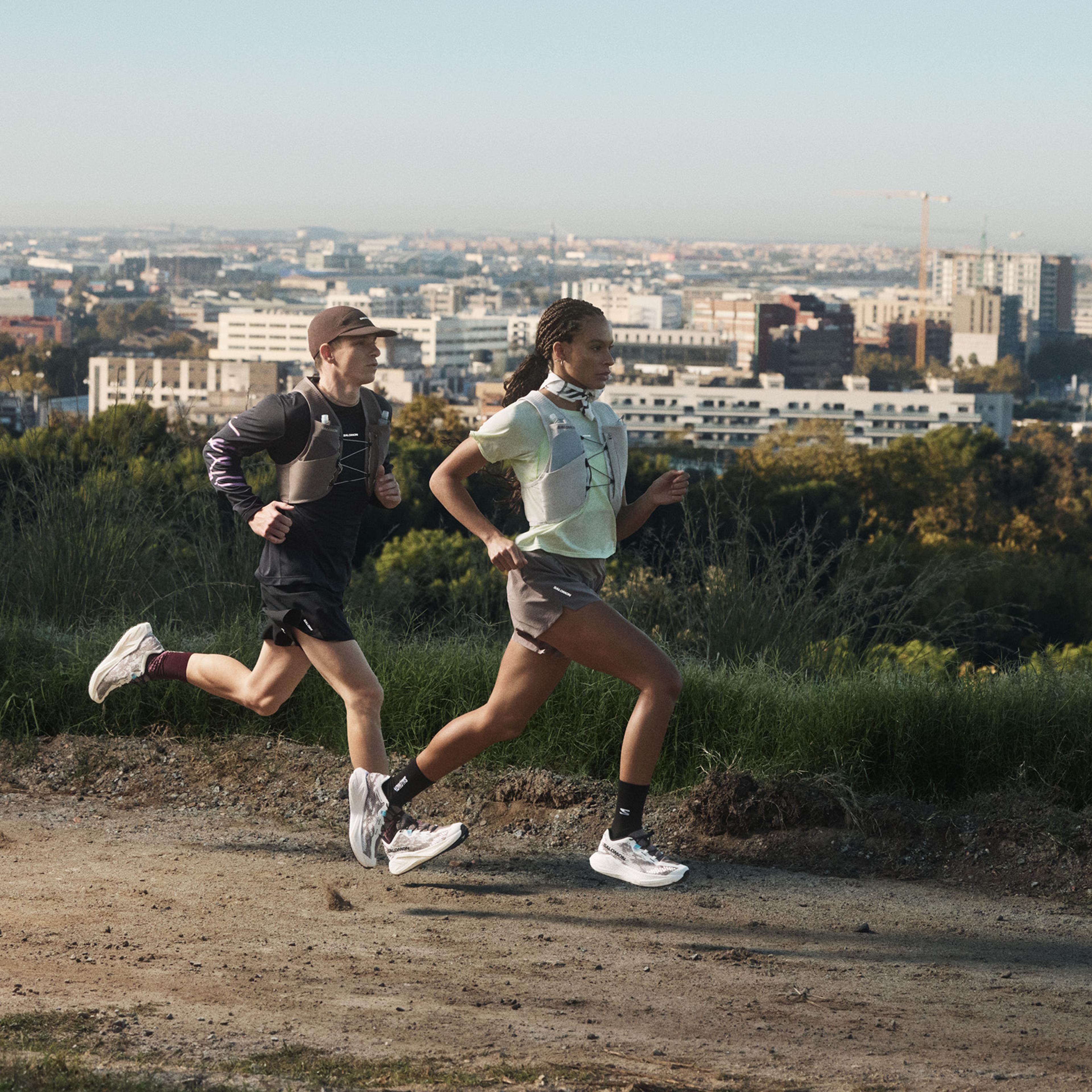 A man and woman trail running with packs on a dirt path overlooking a city.