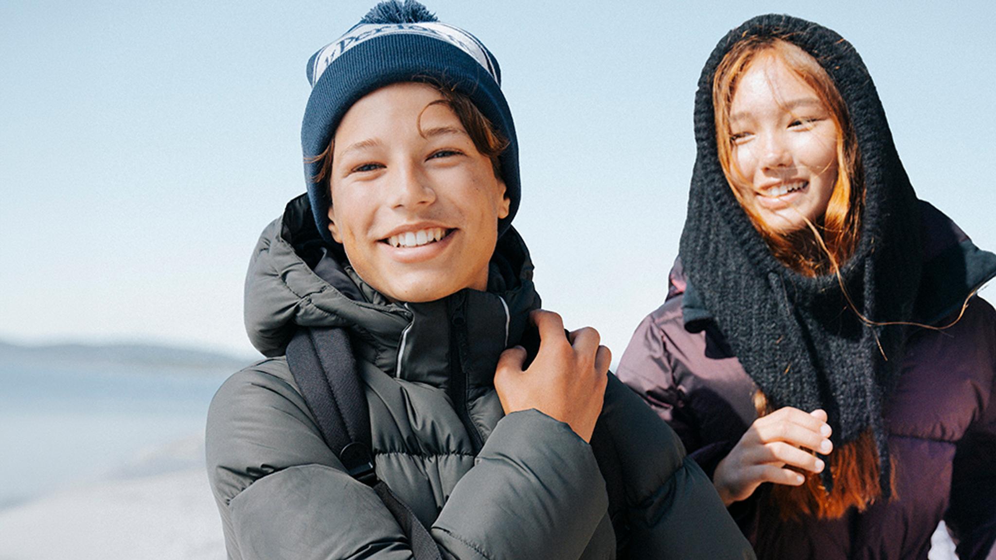 A smiling boy in a blue beanie and dark puffer jacket, with a smiling girl behind him.