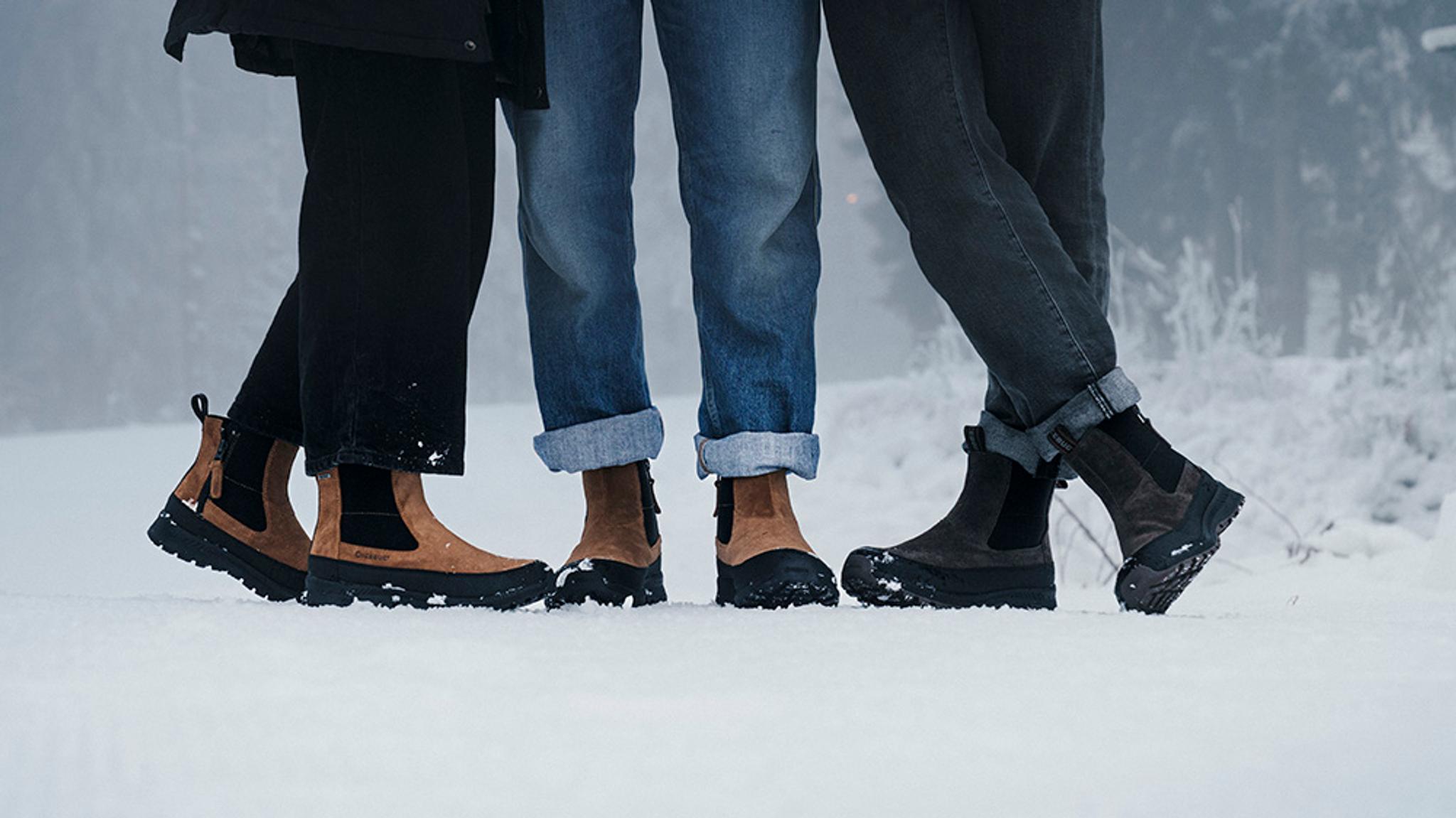 Three people wearing tan and dark grey winter boots stand in the snow.