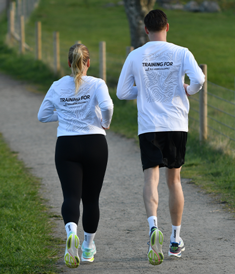 Two people seen from behind, wearing matching white shirts with a topographic map design and "TRAINING FOR" text, jogging on a dirt path.