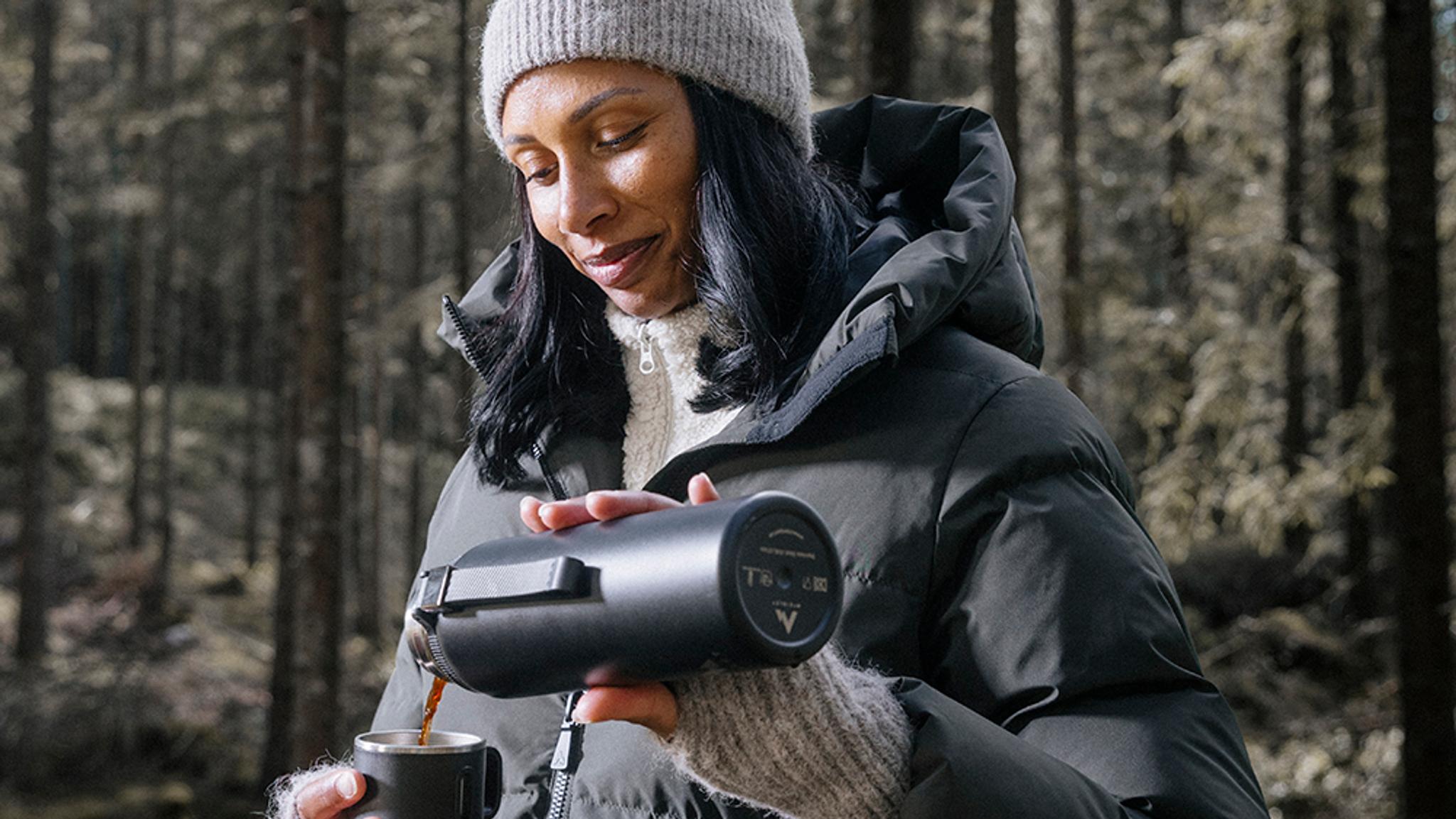 a woman is wearing a jacket and holding a cup of coffee in the forest .