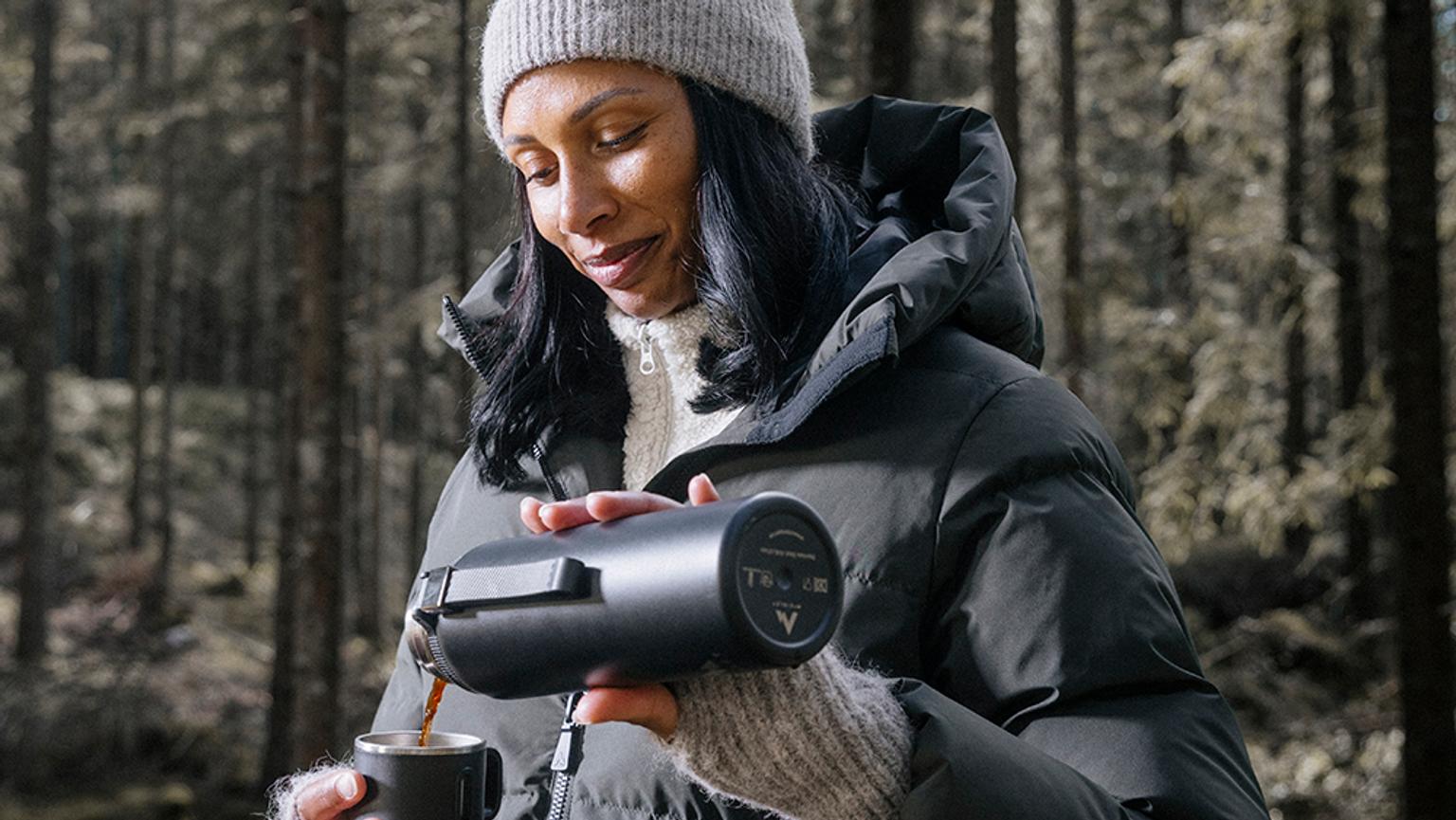 a woman is wearing a jacket and holding a cup of coffee in the forest .