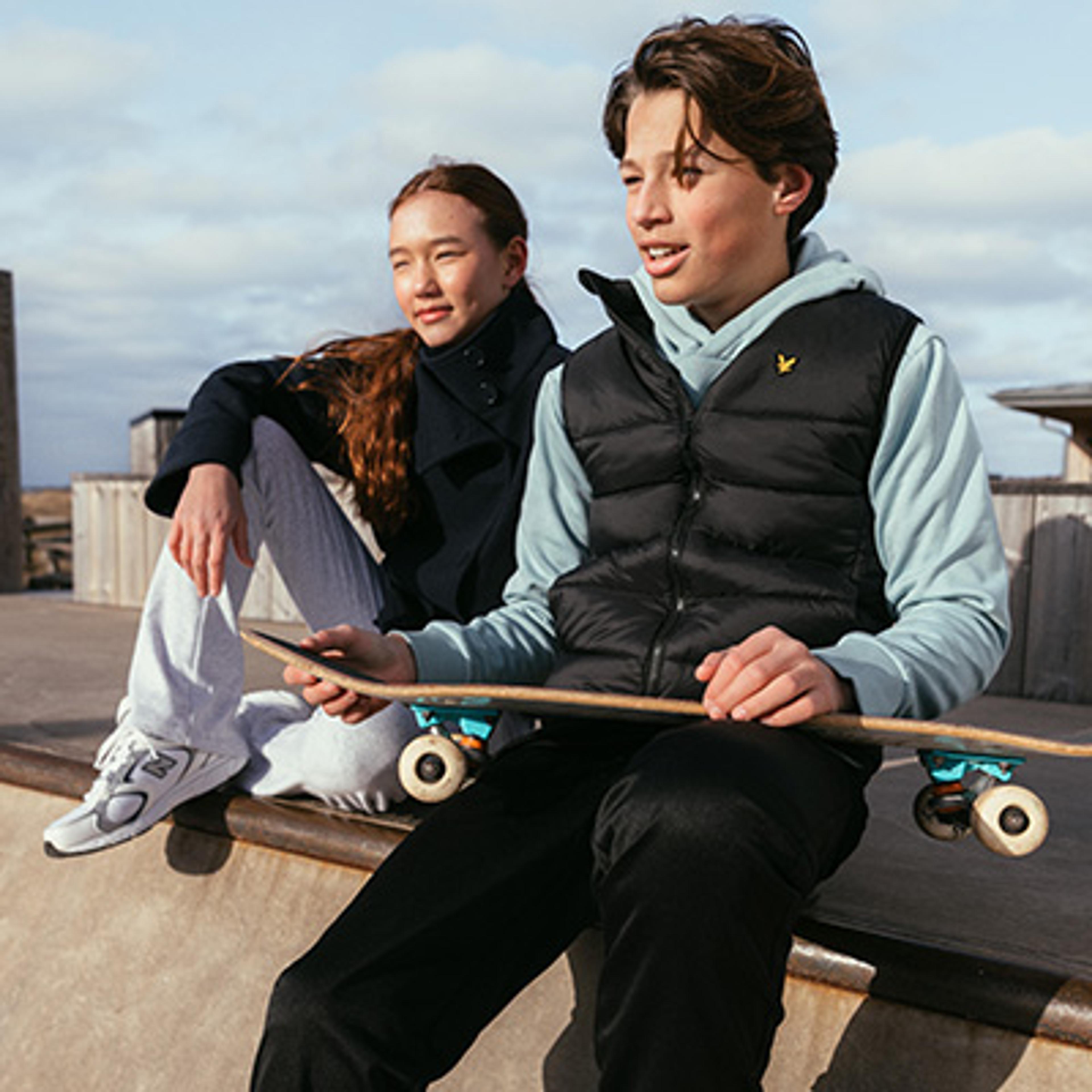 Two teenagers sit at a skate park, the boy in front holding a skateboard.