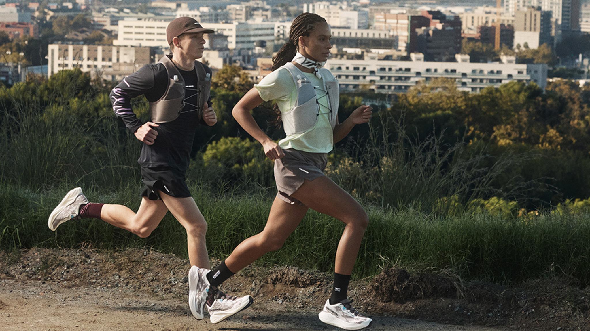 A man and woman running on a dirt trail, both wearing running vests, with a city skyline behind them.